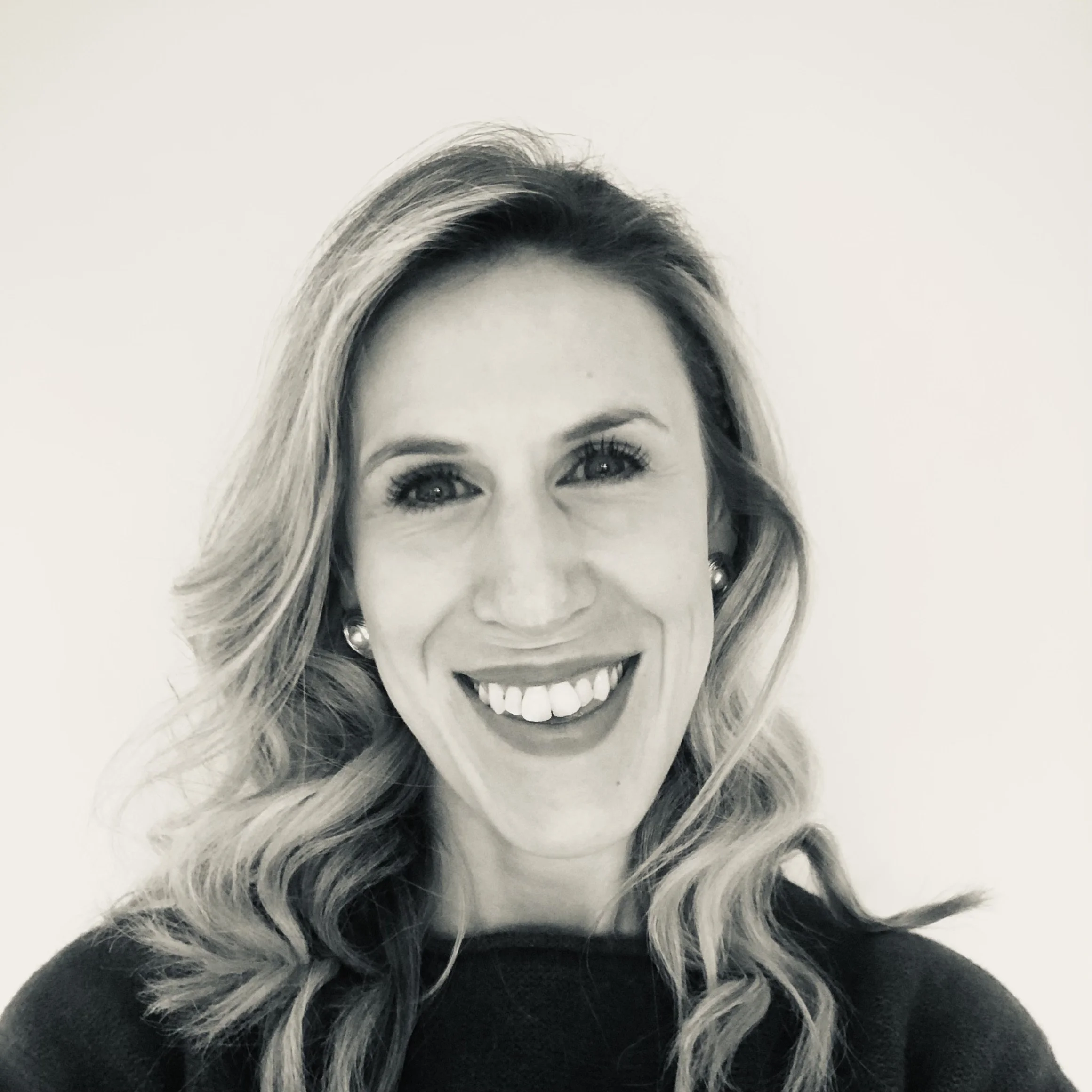 Black and white photo of a smiling woman with wavy shoulder-length hair, wearing pearl earrings and a dark top