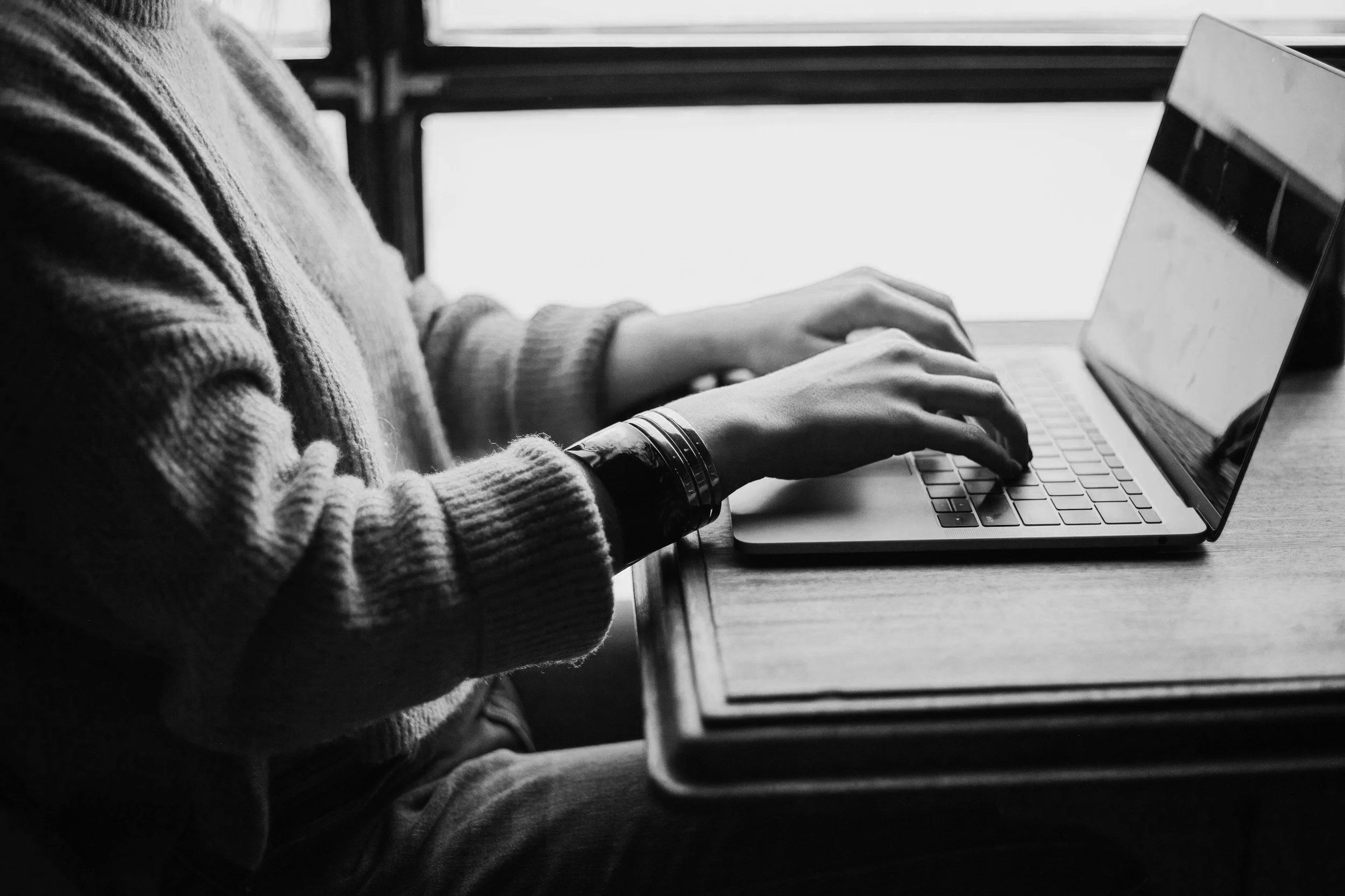 Person wearing a knit sweater typing on a laptop at a desk near a window.