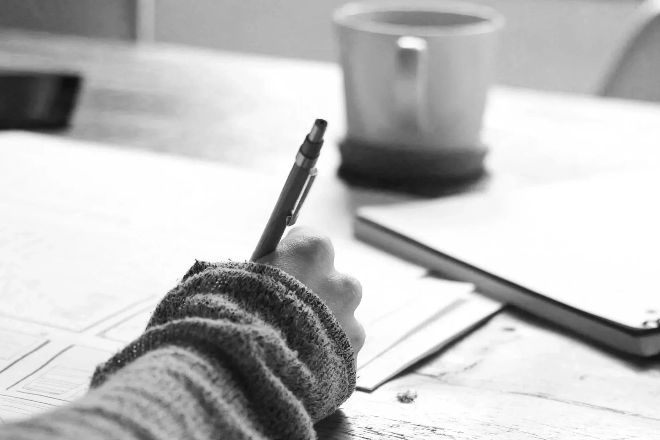 A person wearing a striped sweater is writing with a pen on a piece of paper at a desk. In the background, there is a cup and a closed notebook.