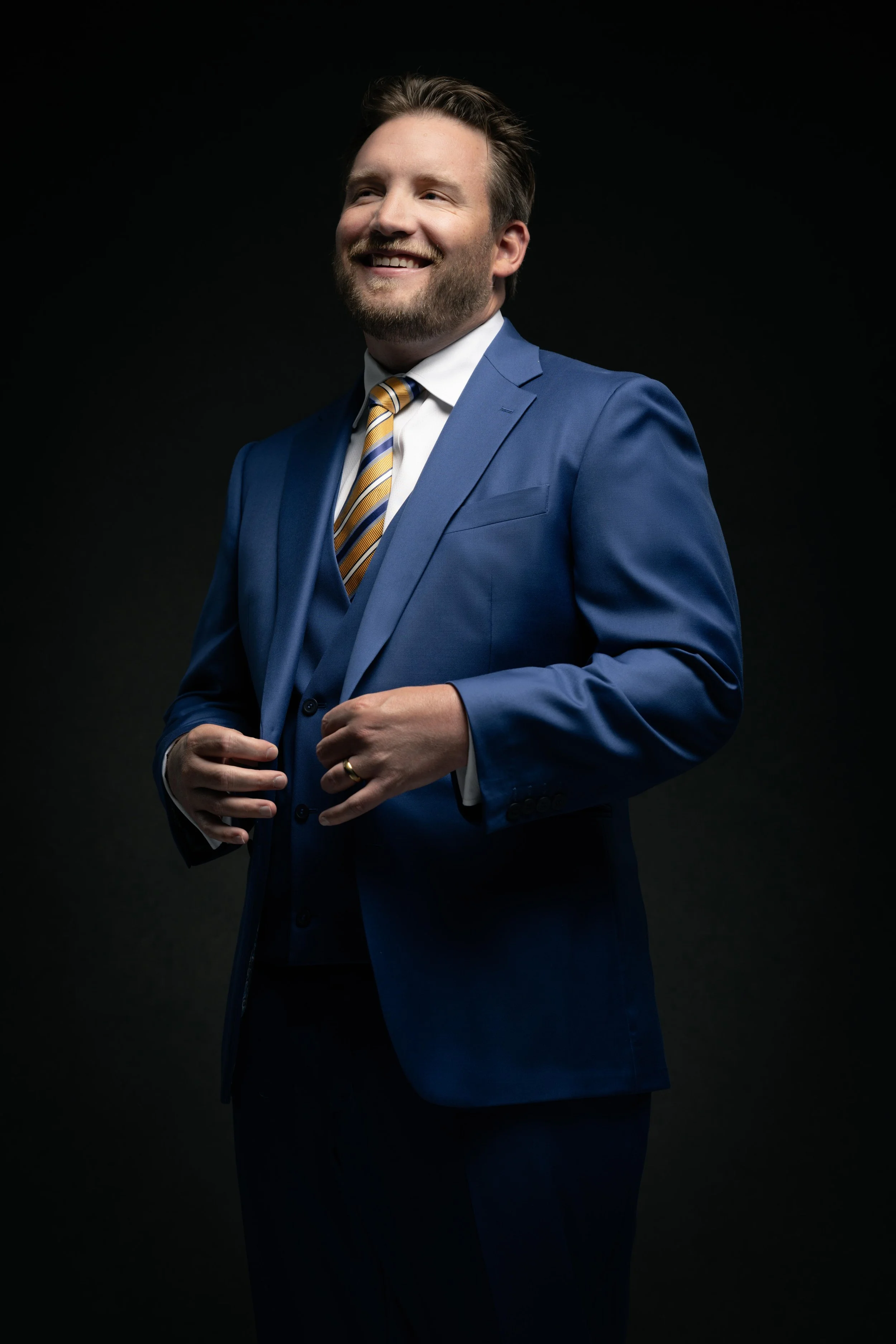 A smiling man with a beard and mustache wearing a blue suit, white shirt, and striped yellow and blue tie, standing against a black background.