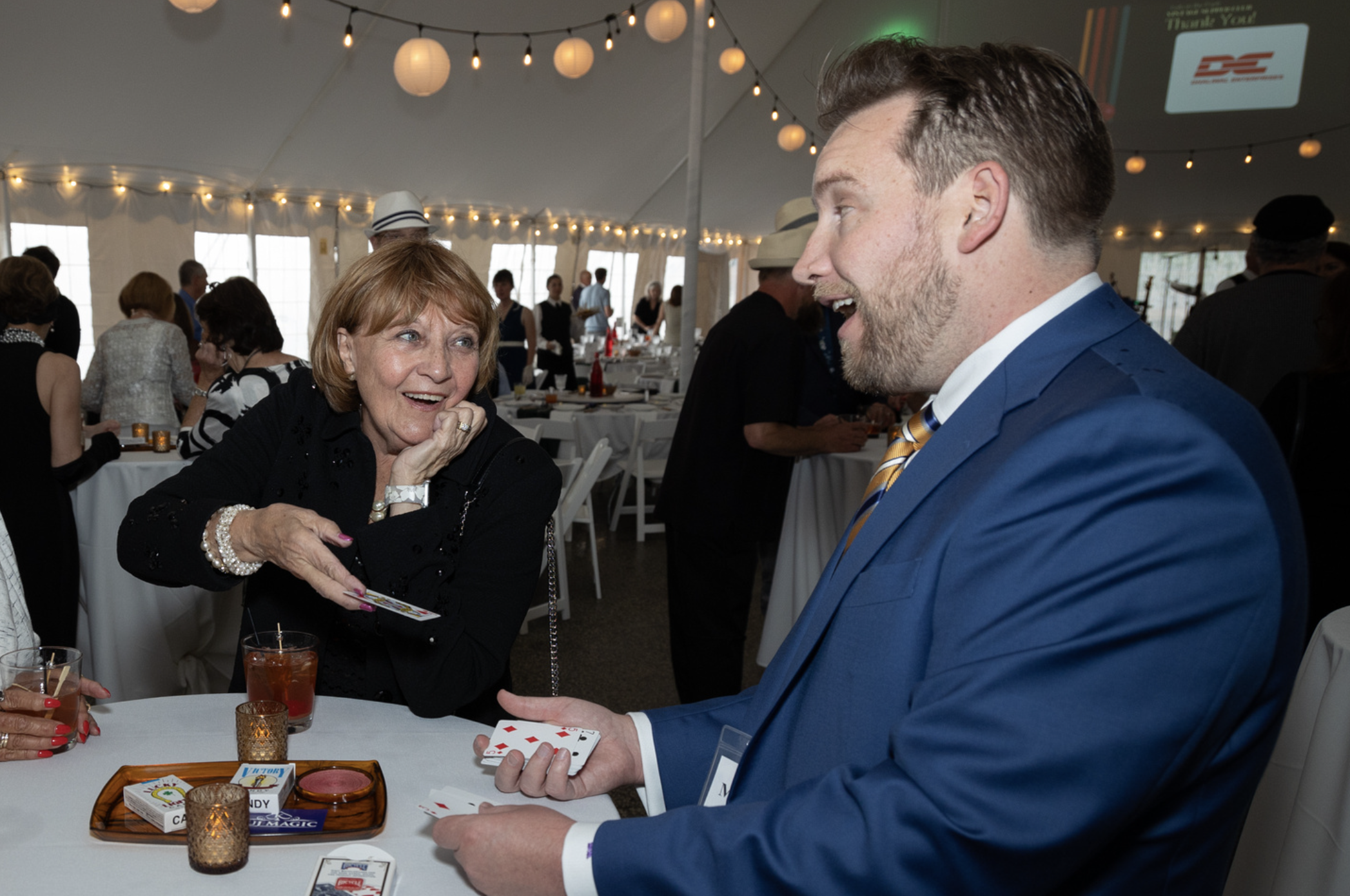 A woman with light brown hair, wearing a black top, is smiling and playing cards with a man in a blue suit, who is showing the cards in his hand. They are at a round table with a card game, drinks, and small lit candle holders. The setting is inside 