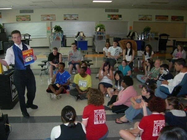 A group of students gathered in a cafeteria, seated on the floor and on chairs, watching a person standing and holding an open book.