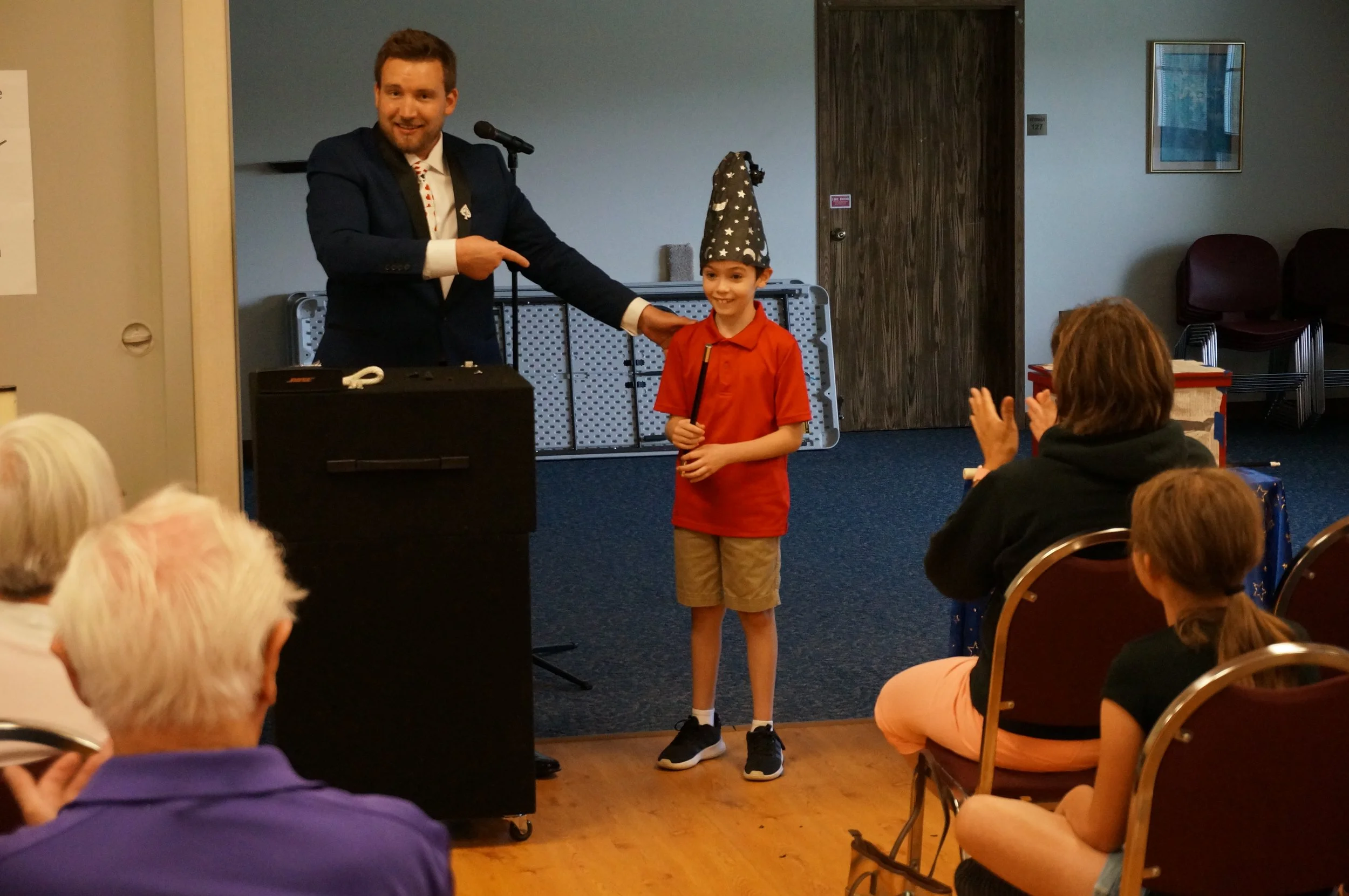 Magician pointing to a boy wearing a wizard hat and holding a magic wand during a performance; audience members clapping.