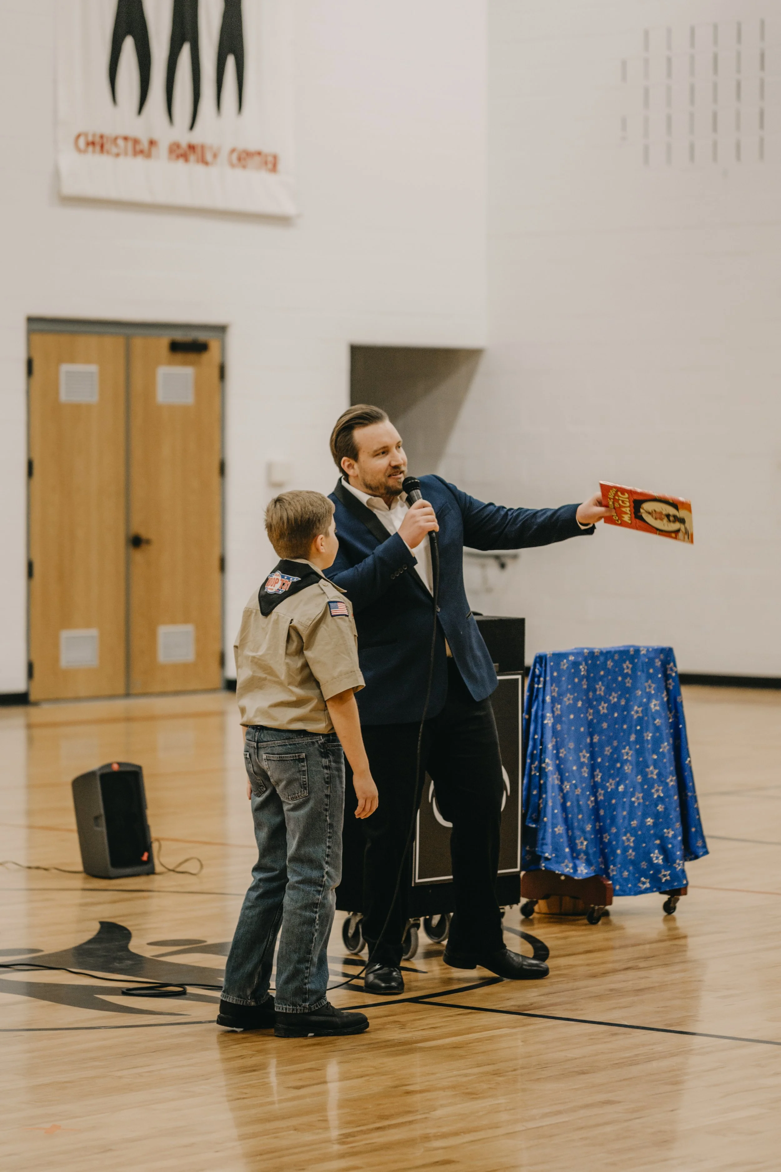 A man in a suit holding a microphone and a book stands next to a boy wearing a scout uniform on a gym floor. There’s a speaker on the ground and a table covered with a blue star-patterned cloth nearby.