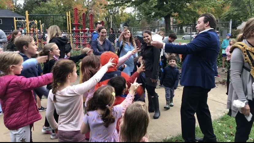 A group of children eagerly reaching towards a man in a blue suit at a playground. Parents and children are gathered around, some taking photos with smartphones. The playground equipment is visible in the background, surrounded by trees.
