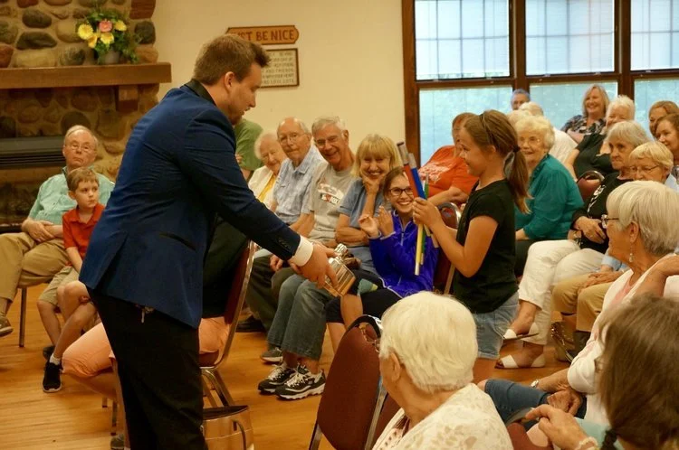 A magician in a blue suit performs a trick for a young girl holding colorful scarves, in front of an audience of seated adults and children in a community room.