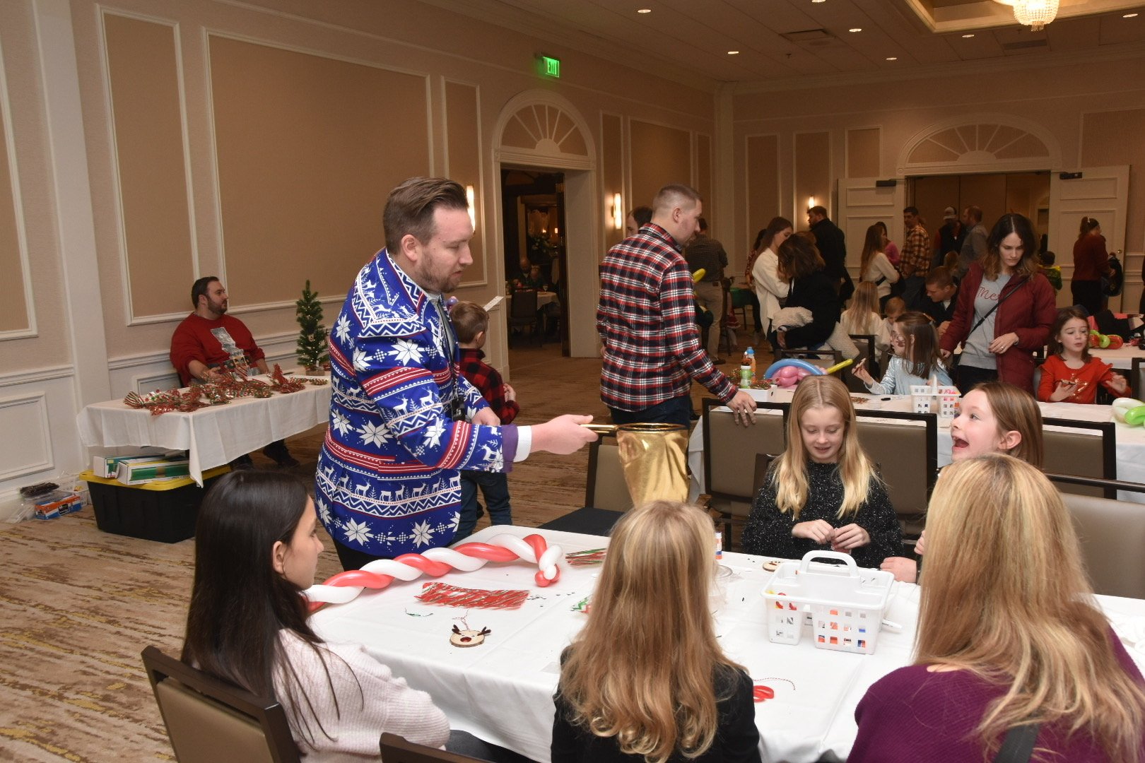 A festive gathering in a decorated room with children and adults participating in holiday activities at a table. A man is wearing a blue holiday-themed sweater and engaging with the children. Various craft materials are on the table.