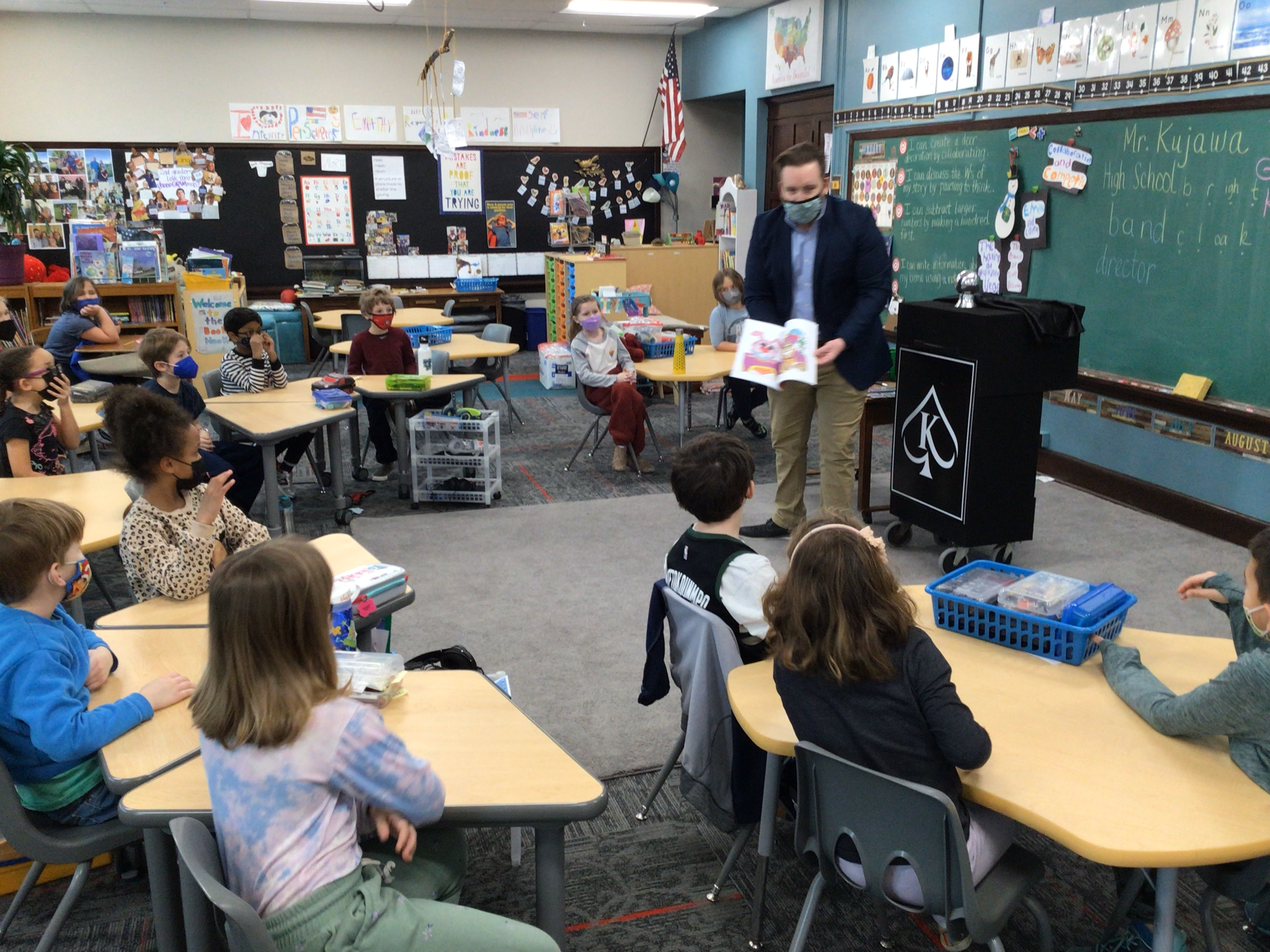 A classroom with students seated at desks and a teacher standing in front of a chalkboard, holding a book. Some students are wearing masks. The room is decorated with educational materials and posters. The teacher is showing the book to the class.
