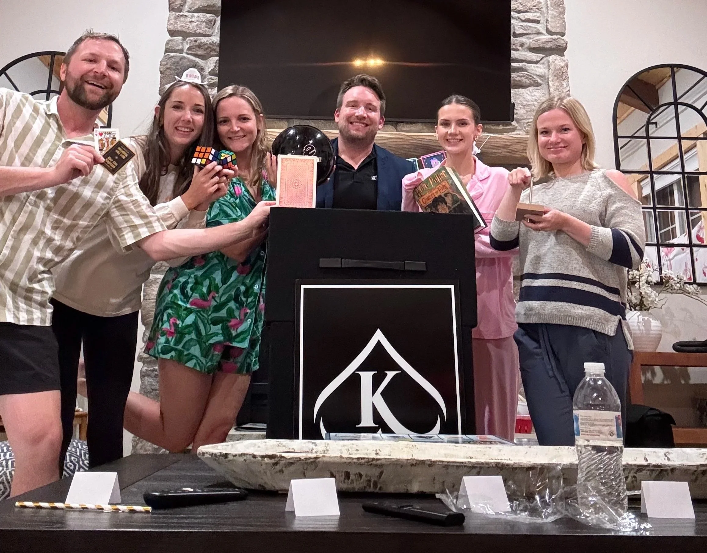 Group of people celebrating at a party or gathering, standing behind a table with a large black box featuring a white logo, possibly a card or game, in a room with a stone fireplace and wood-paneled ceiling.