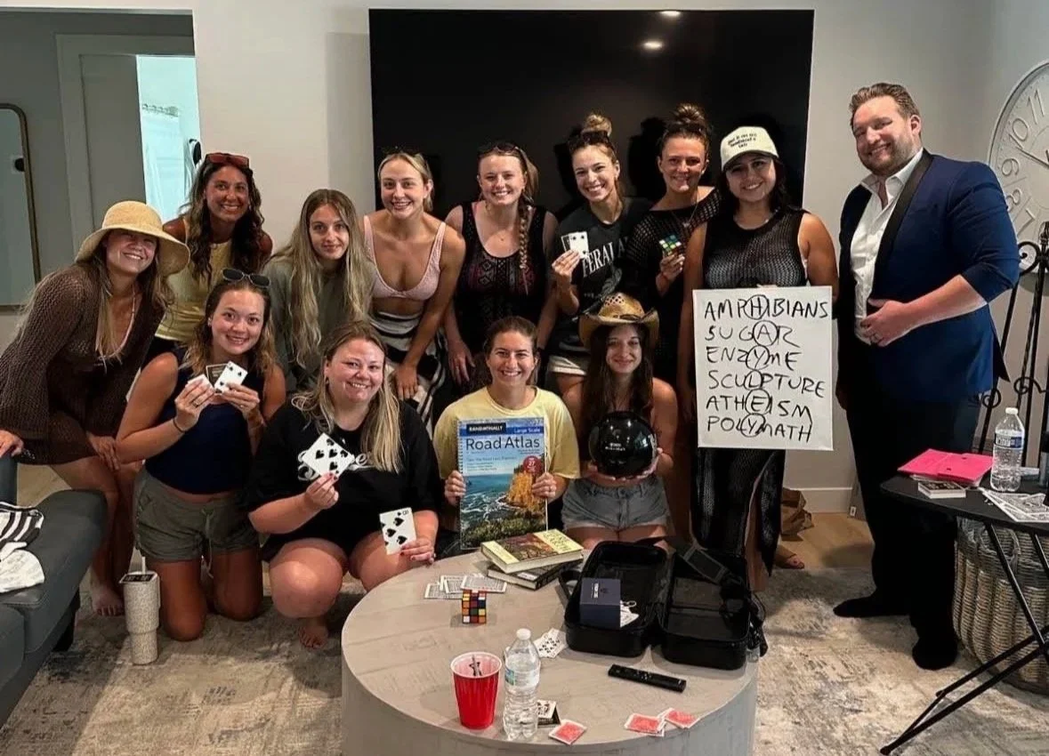Group of women and one man gathered in a living room, smiling and holding playing cards, a Rubik's Cube, and a black helmet, with a large TV and a sign with words in the background.