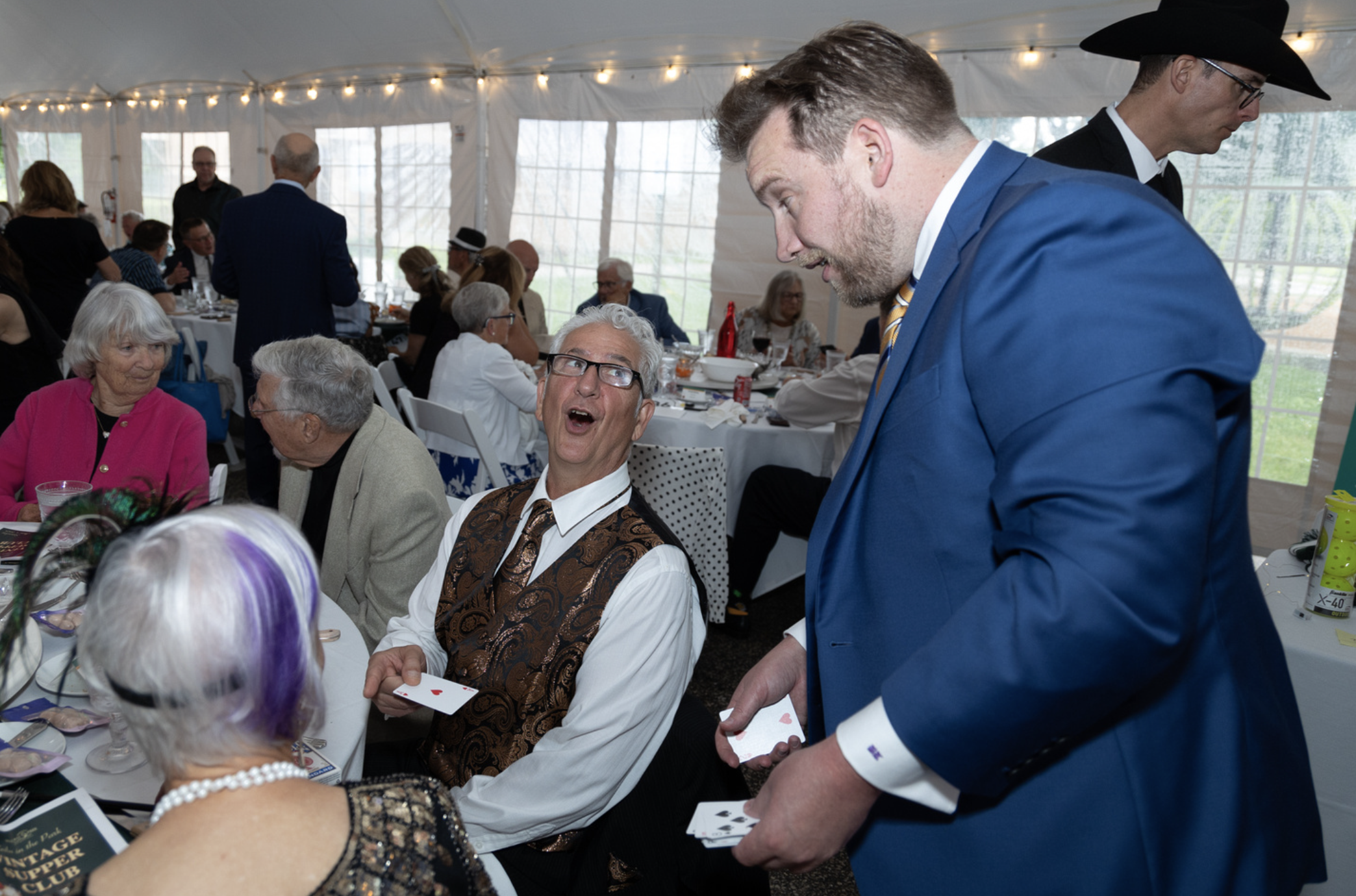 People playing cards at a banquet, with two men in suits and hats engaging in conversation, under a decorated tent.