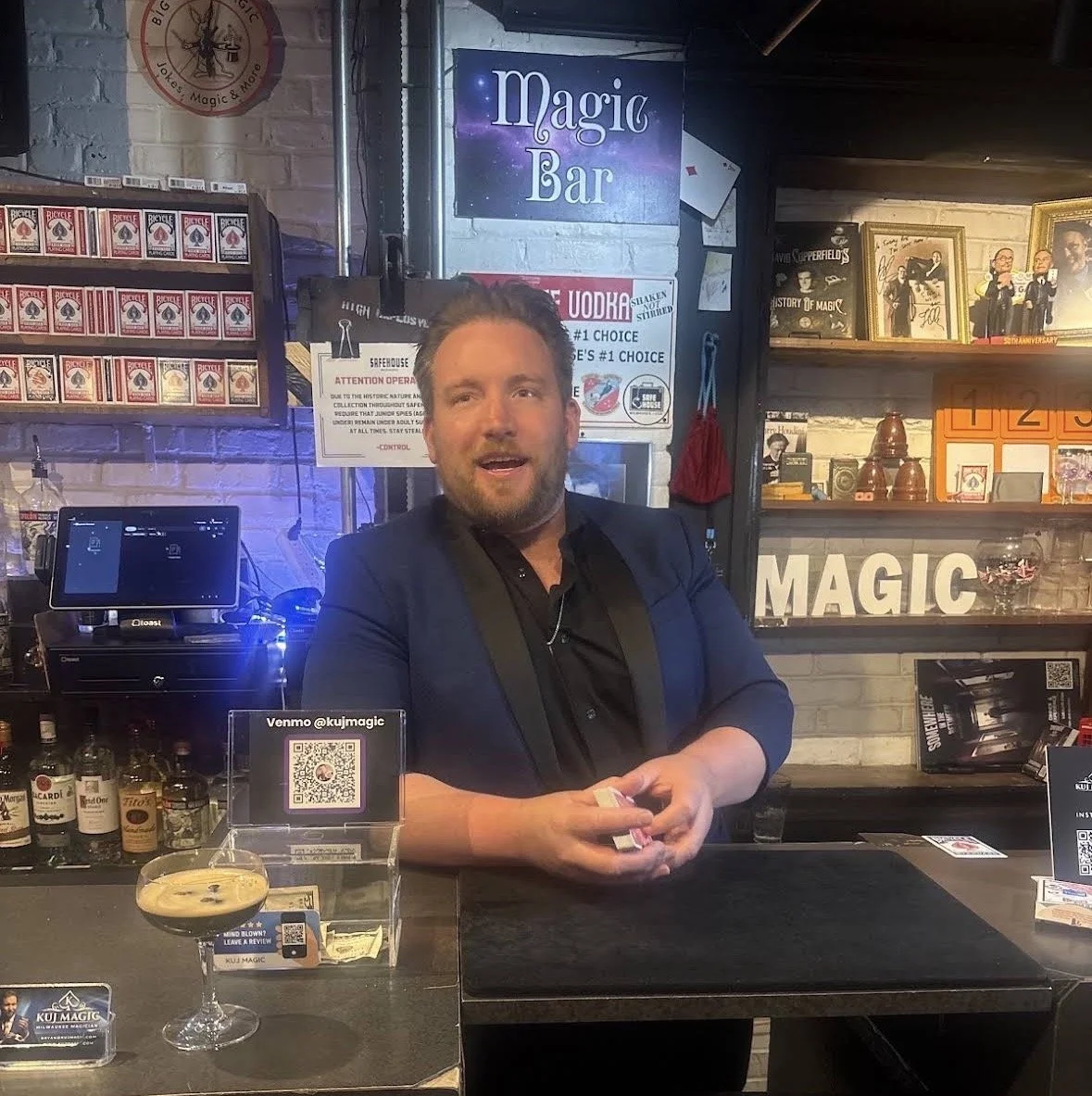 A man standing behind a bar counter at a magic bar, holding cards in his hands and smiling. The background includes shelves with framed photos, cards, and bottles, with signs for 'Magic Bar' and a QR code for Venmo.