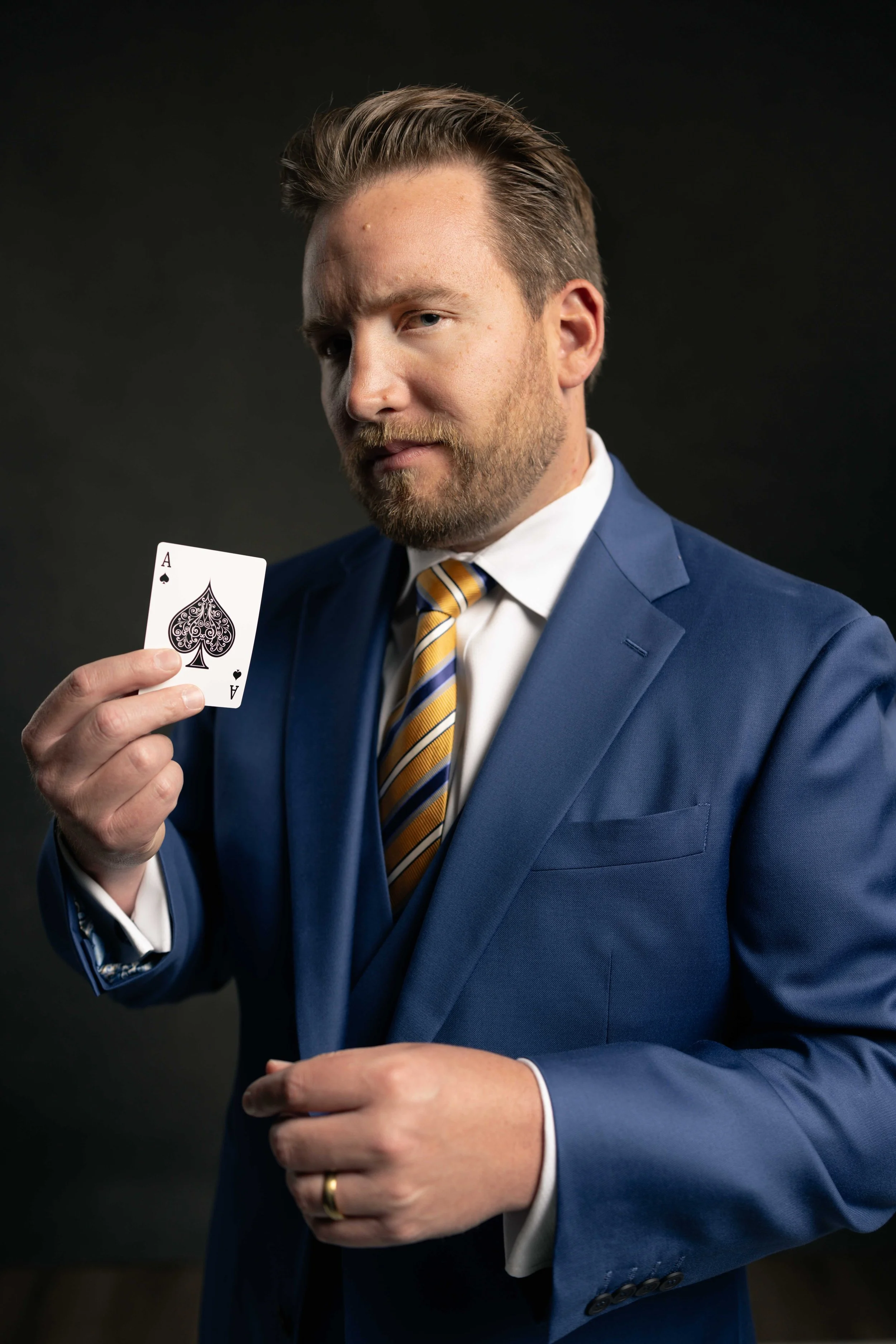 A man in a blue suit with a white shirt and yellow striped tie holding an ace of spades playing card in his right hand.