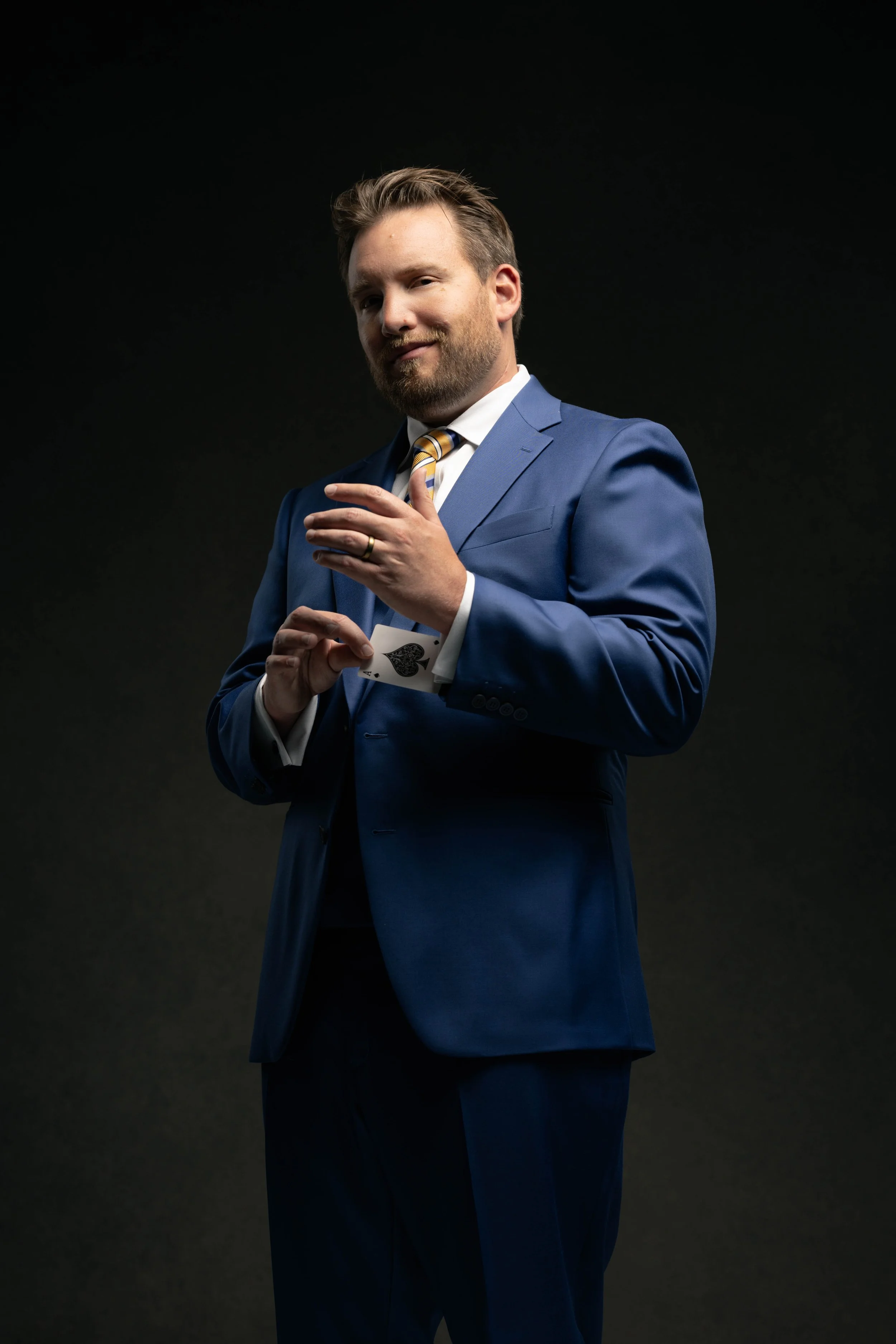 A man wearing a blue suit with a striped tie holding a playing card, the Ace of Spades, against a black background