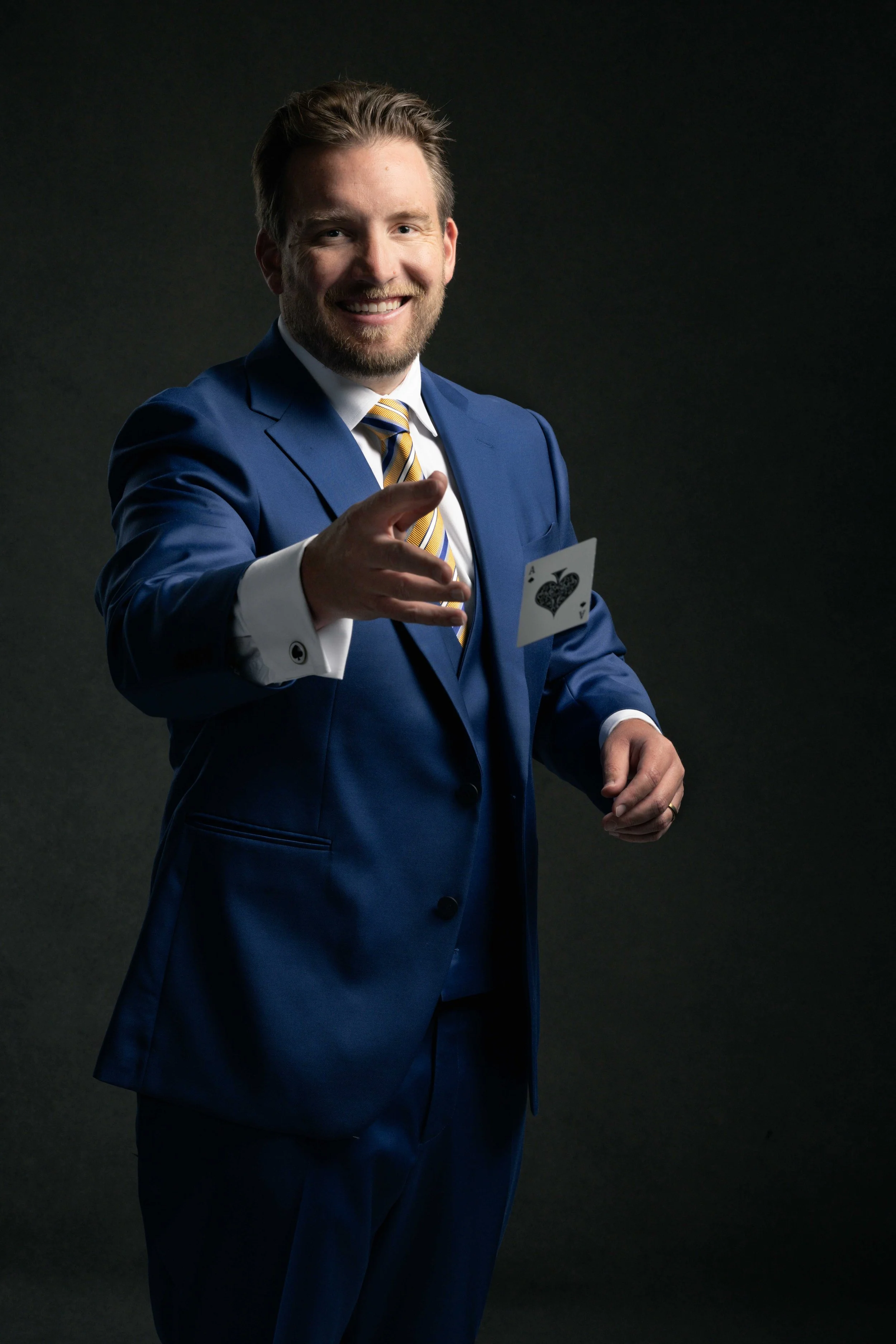 A man in a blue suit with a striped yellow and blue tie, smiling, holding a card with a spade symbol, pointing at the camera against a dark background.