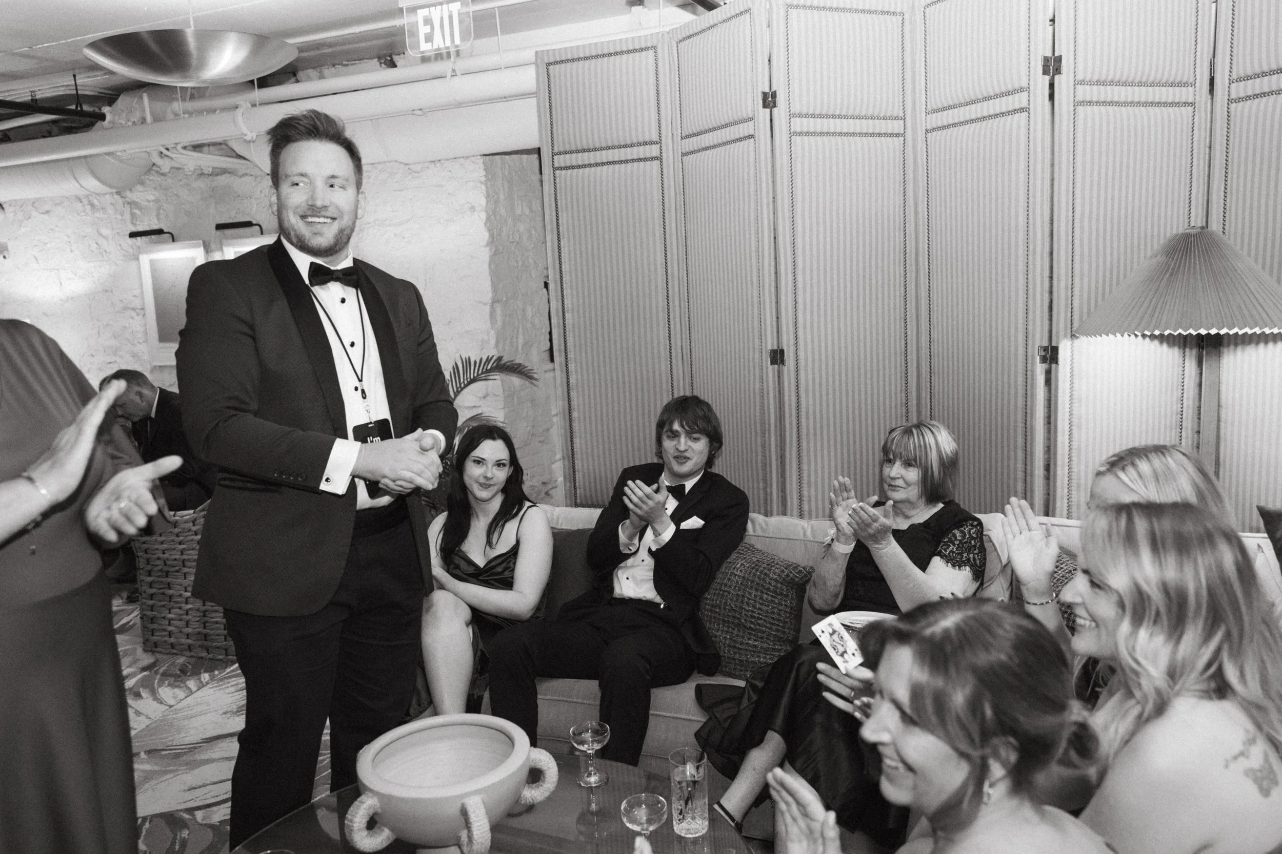 A man in a tuxedo shaking hands at a social gathering, with several seated women clapping and smiling around him in an indoor setting.