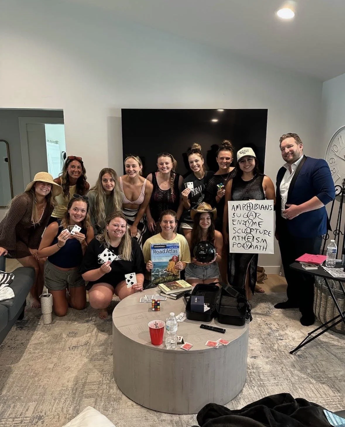 Group of people gathered indoors, holding playing cards, Rubik's cube, and a black helmet, with a whiteboard displaying words. There are books, a magazine, water bottles, and various items on a round coffee table in front of them.