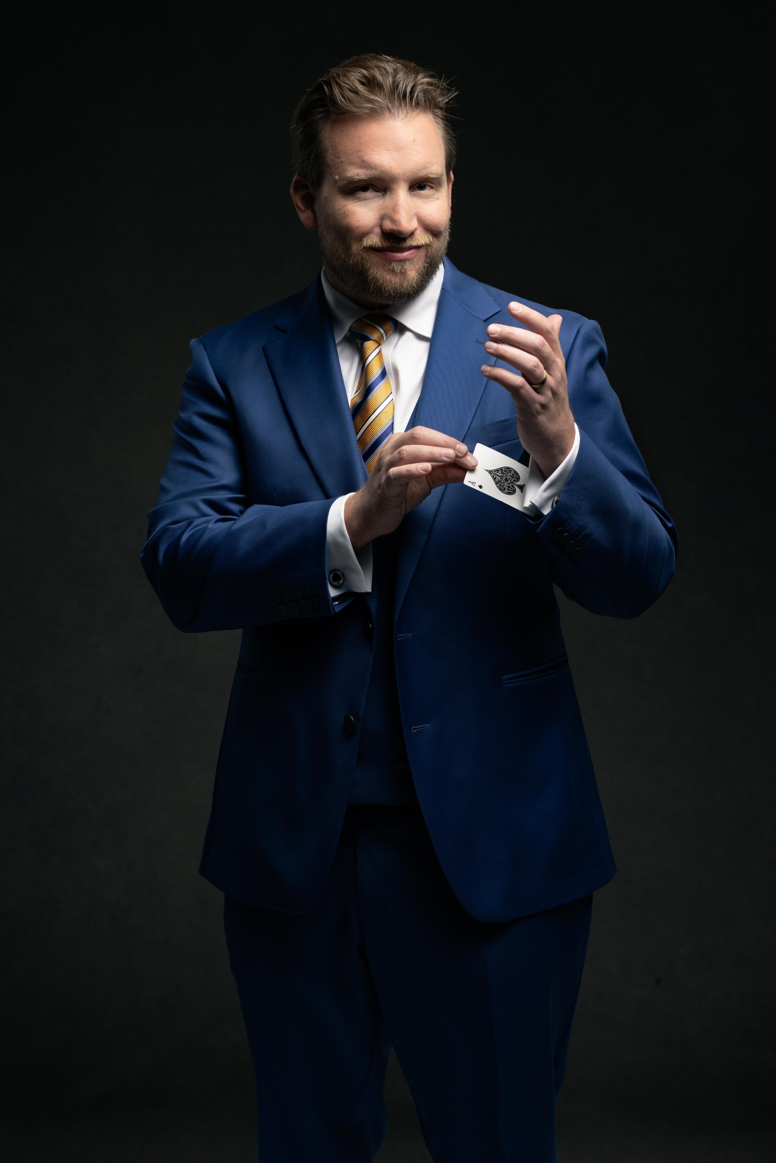 A man in a blue suit with a striped tie holding a playing card, the ace of spades, against a dark background.