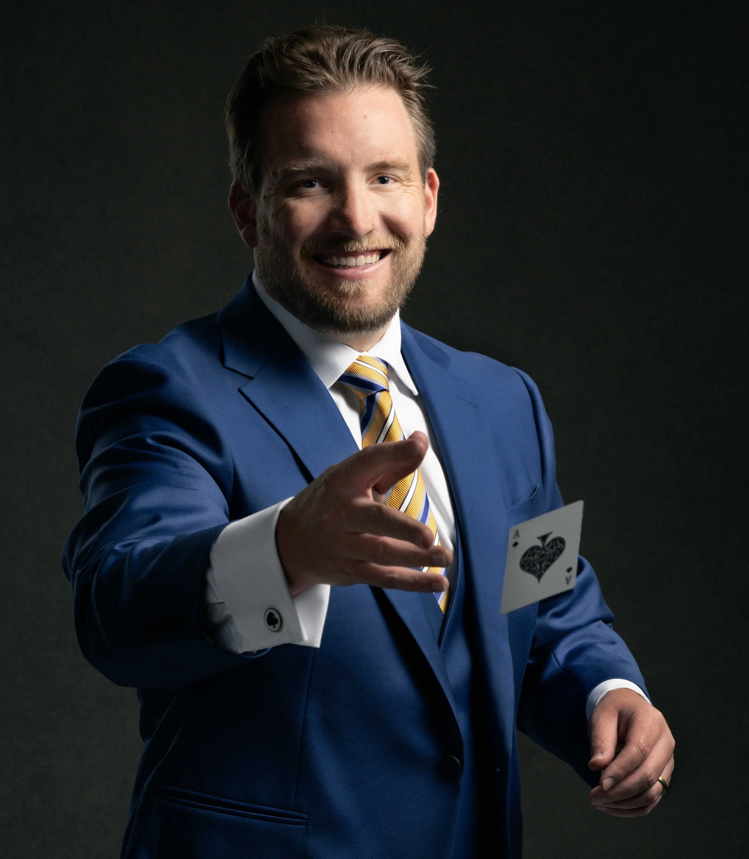 A man in a royal blue suit, white shirt, and yellow striped tie, smiling and pointing forward with his right hand, holding a playing card in his left hand, against a dark background.
