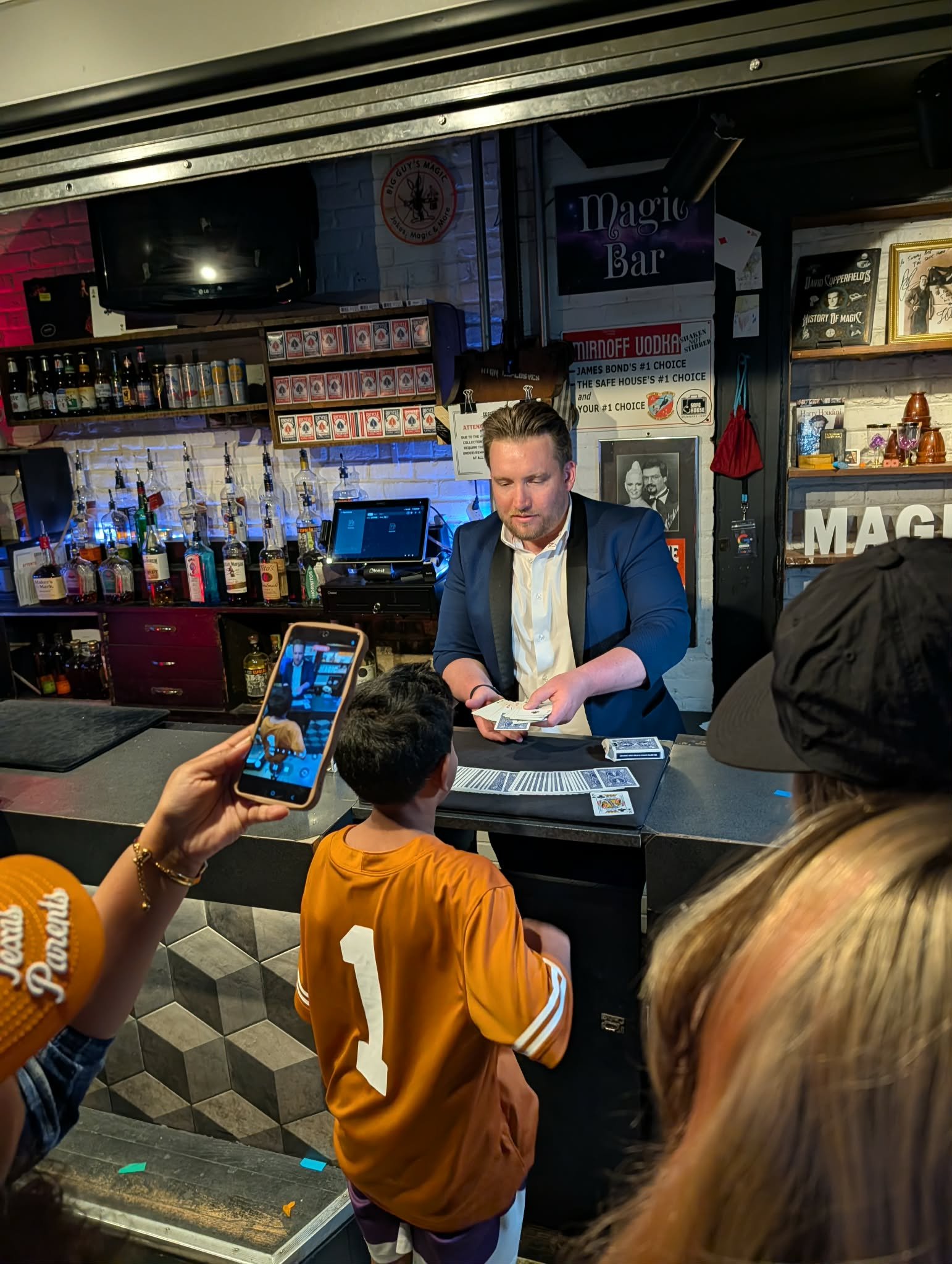 A man in a suit exchanging money with customers at a bar or nightclub. There are people in line, one wearing a football jersey with the number 1. On the wall, signs read 'Magic Bar' and 'MIRNOFF UOAKA,' with bottles of liquor behind the counter and a