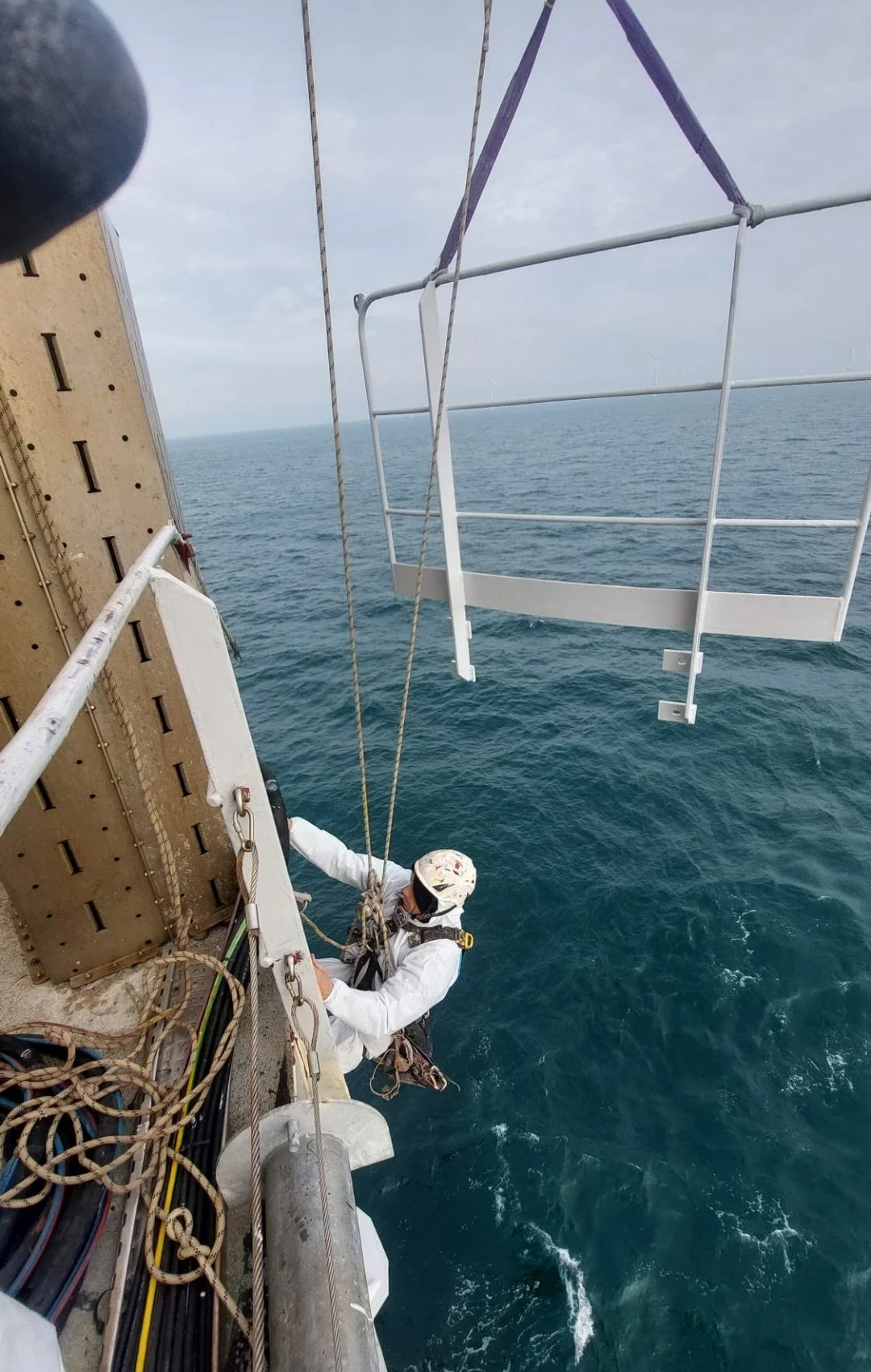 A person in safety gear climbing a metal ladder on the side of a tall structure above the ocean, with a cloudy sky in the background.