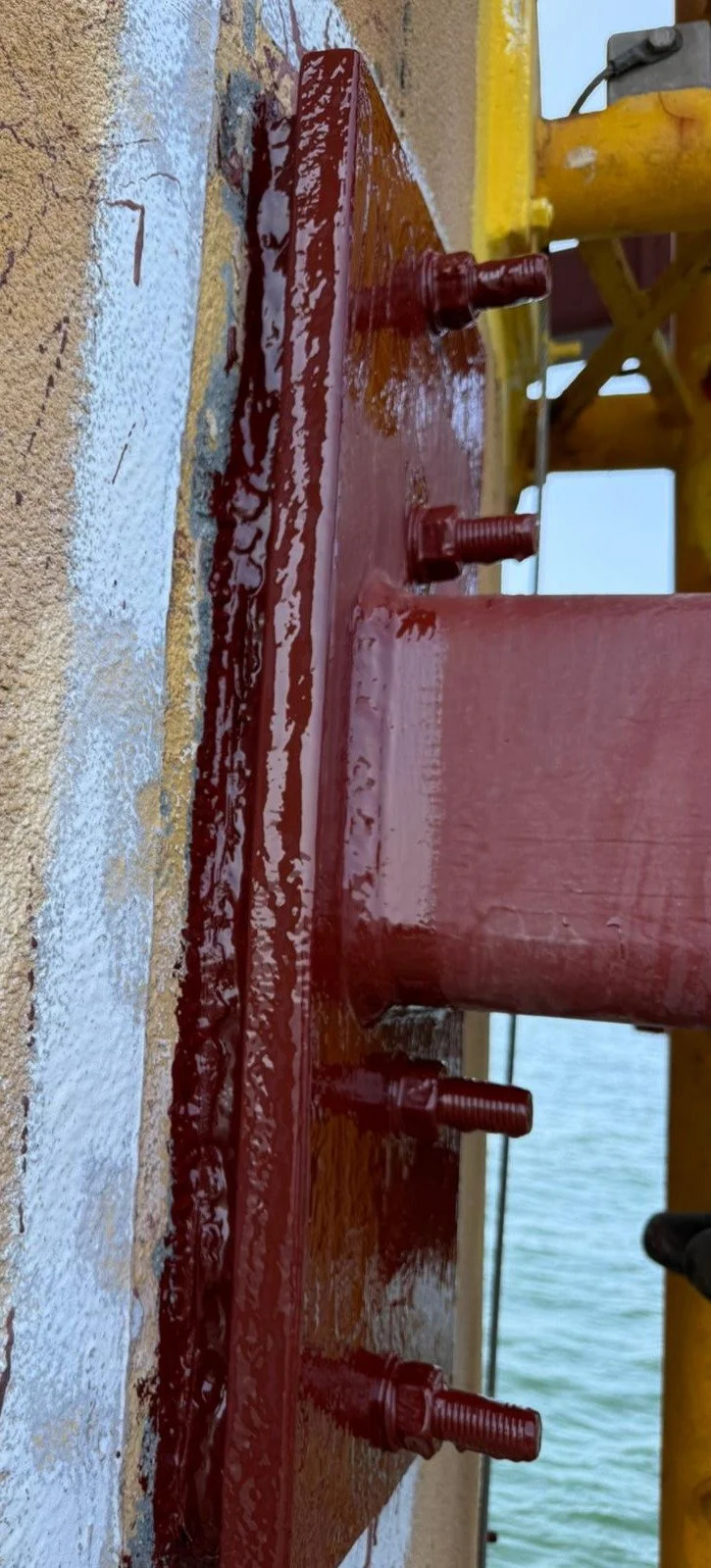 Close-up of a steel pier with red paint, bolts, and a yellow support structure near water.