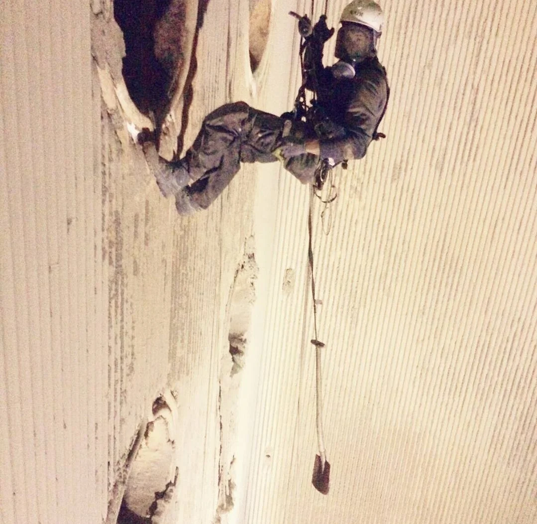 A person in safety gear, including a helmet and harness, is climbing or working on a vertical surface, possibly a wall or a tower.
