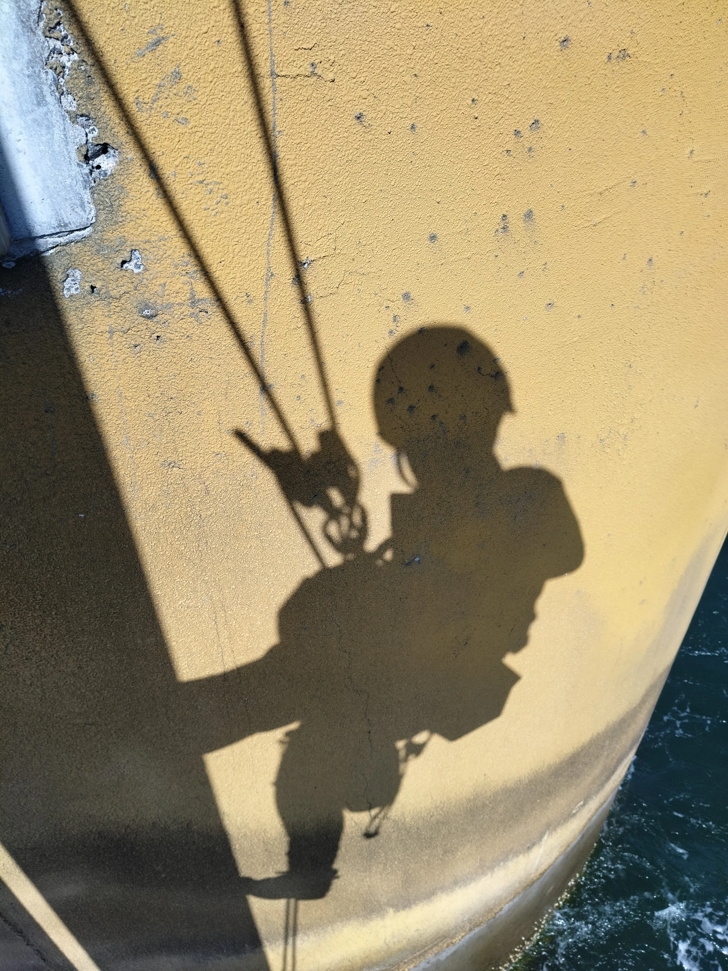 Shadow of a person wearing a helmet on a wall near water, with ropes attached to the helmet.