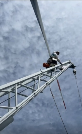 A worker climbing a tall ladder fixed to a wind turbine against a cloudy sky.