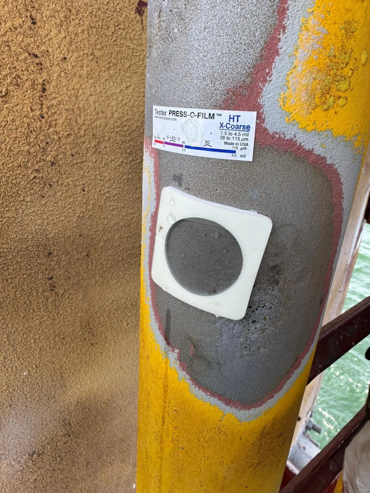 Close-up view of a yellow metal pole on a bridge with a weathered paint and rust patches, and an attached sensor or camera mounted on a white square base.