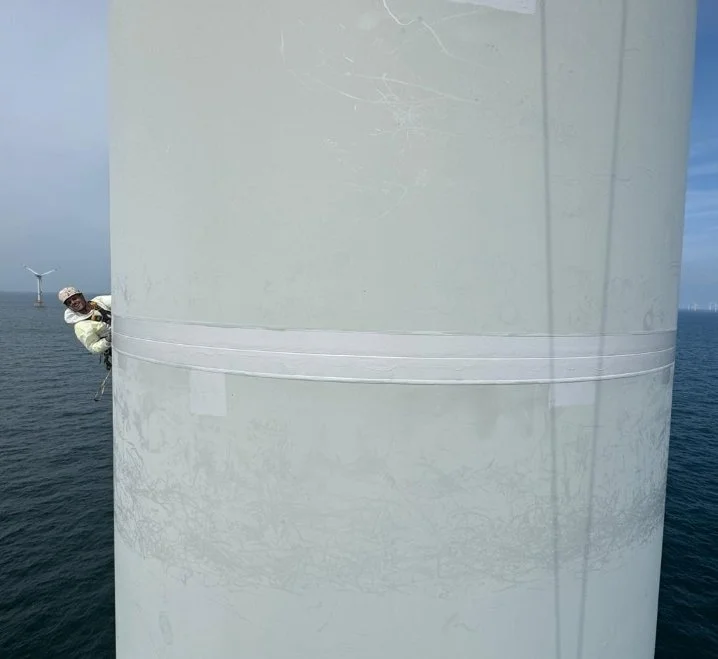 Person climbing or working on a large white wind turbine tower over the ocean.