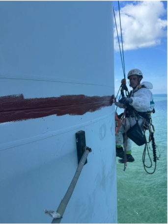 A person wearing safety gear, including a helmet and harness, working on the side of a large white boat with a red stripe, in a green offshore area under a partly cloudy sky.
