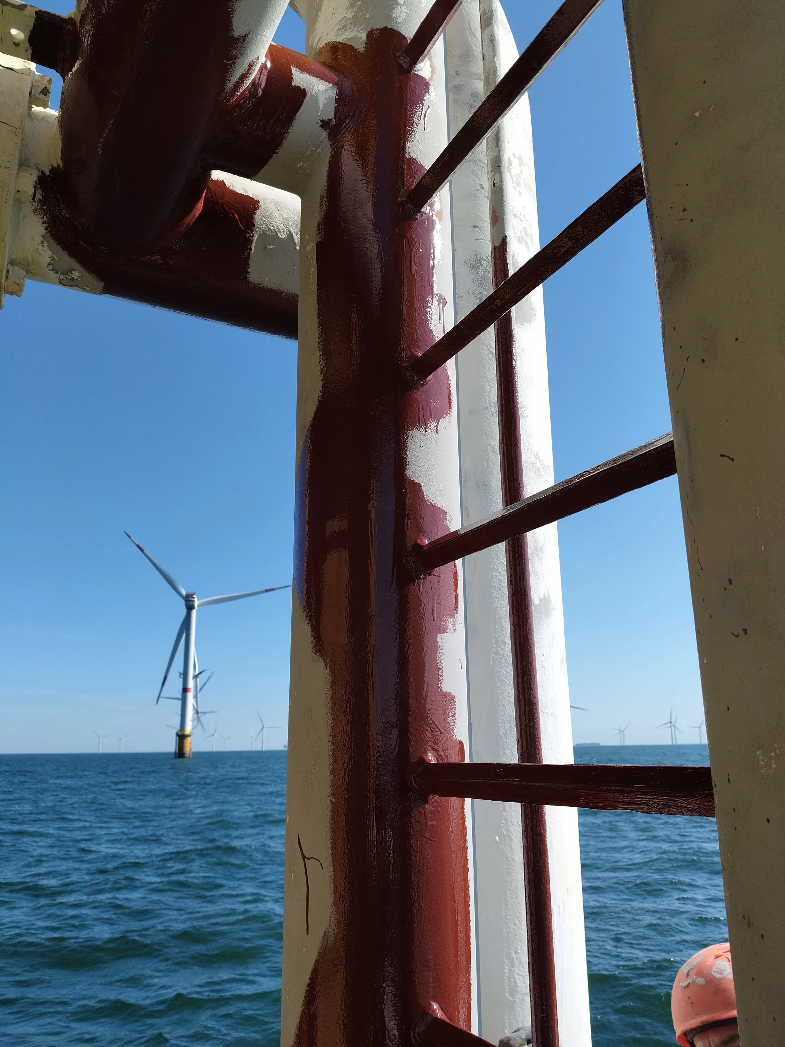 View from a boat showing a red and white pipe structure with rust and fasteners, with offshore wind turbines and blue sky in the background.