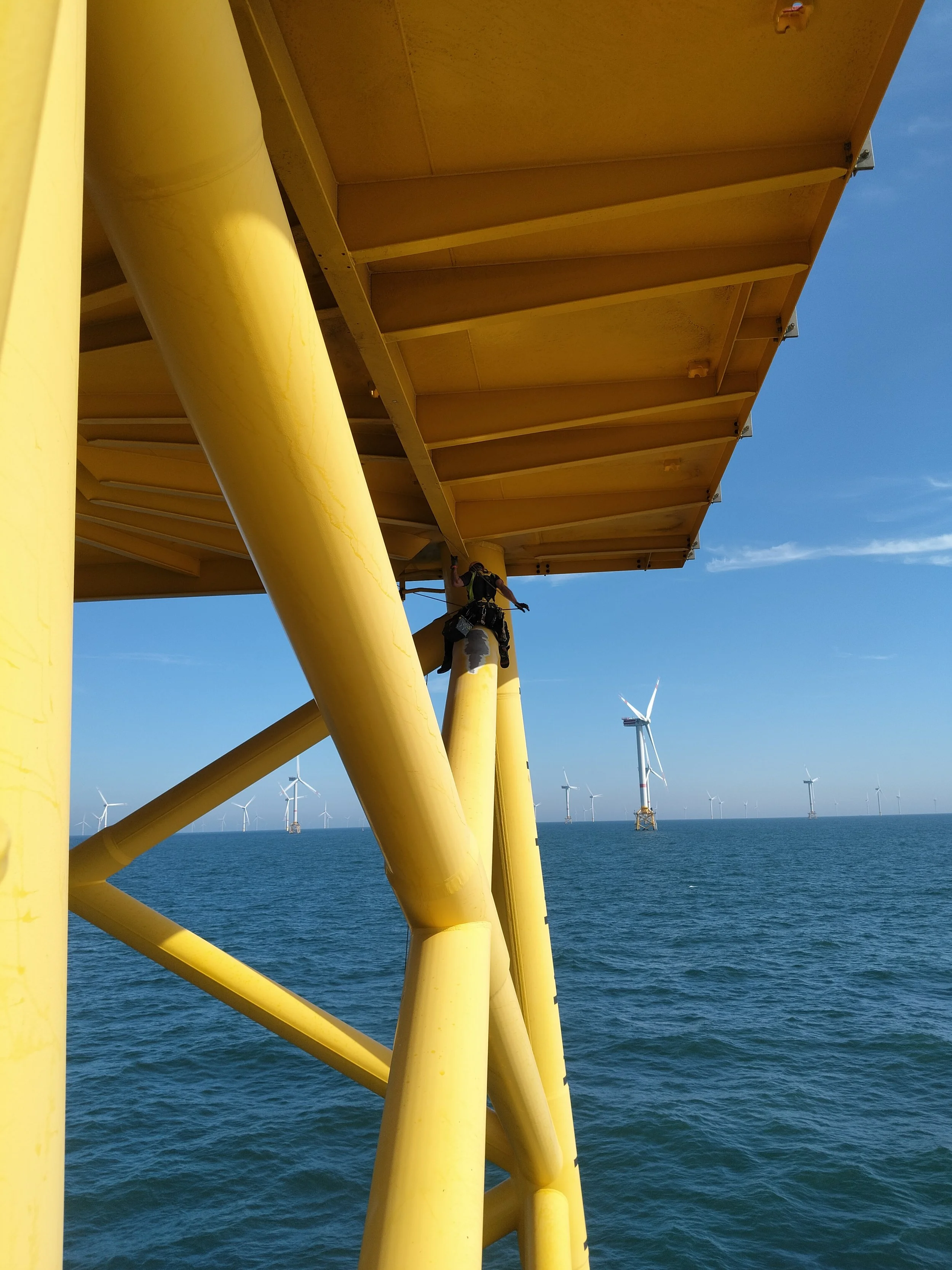 Offshore wind turbines seen from a yellow offshore platform structure in the ocean, under a blue sky.