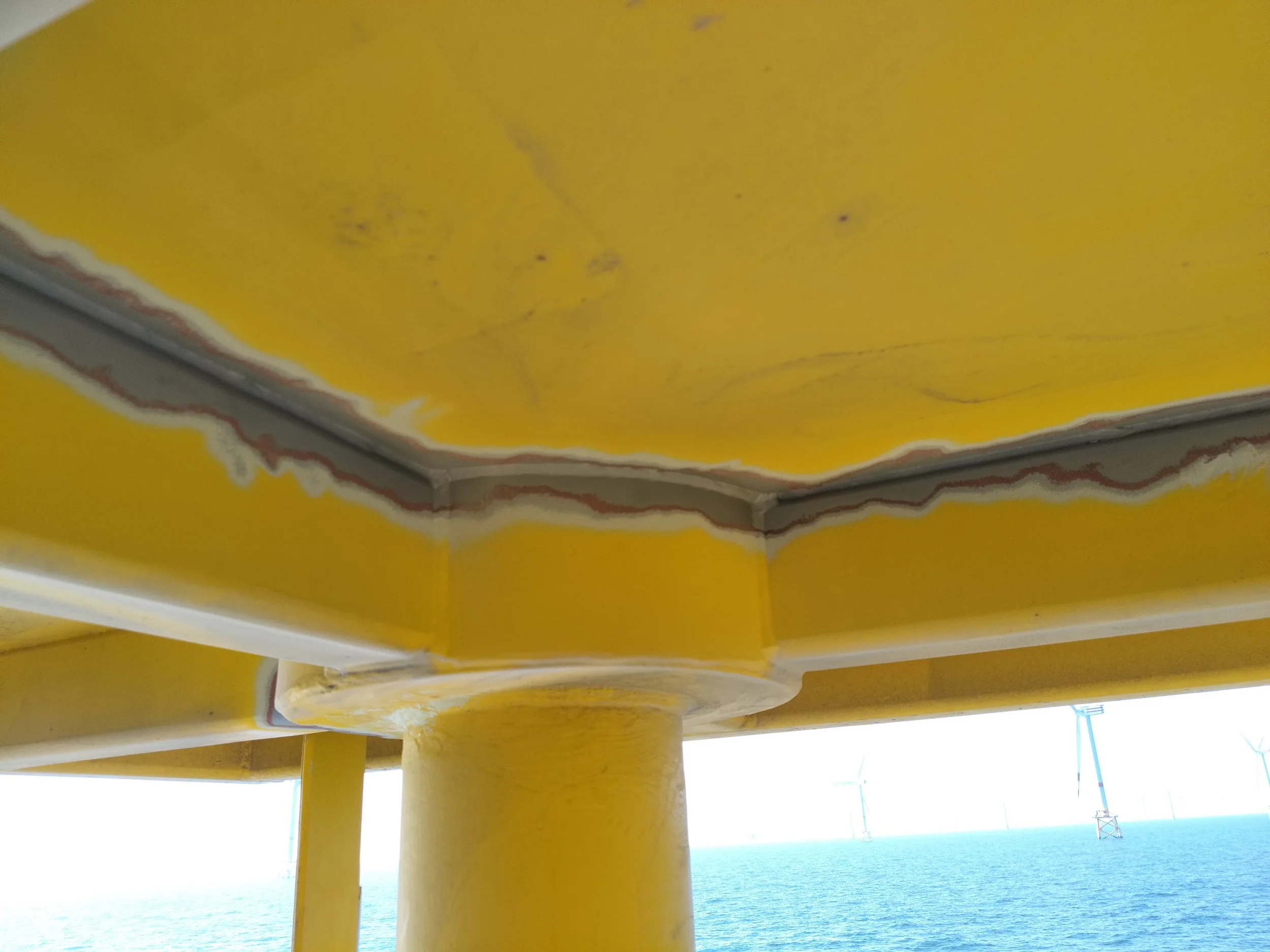 Close-up of the underside of a yellow offshore oil platform with wind turbines in the background over the ocean.