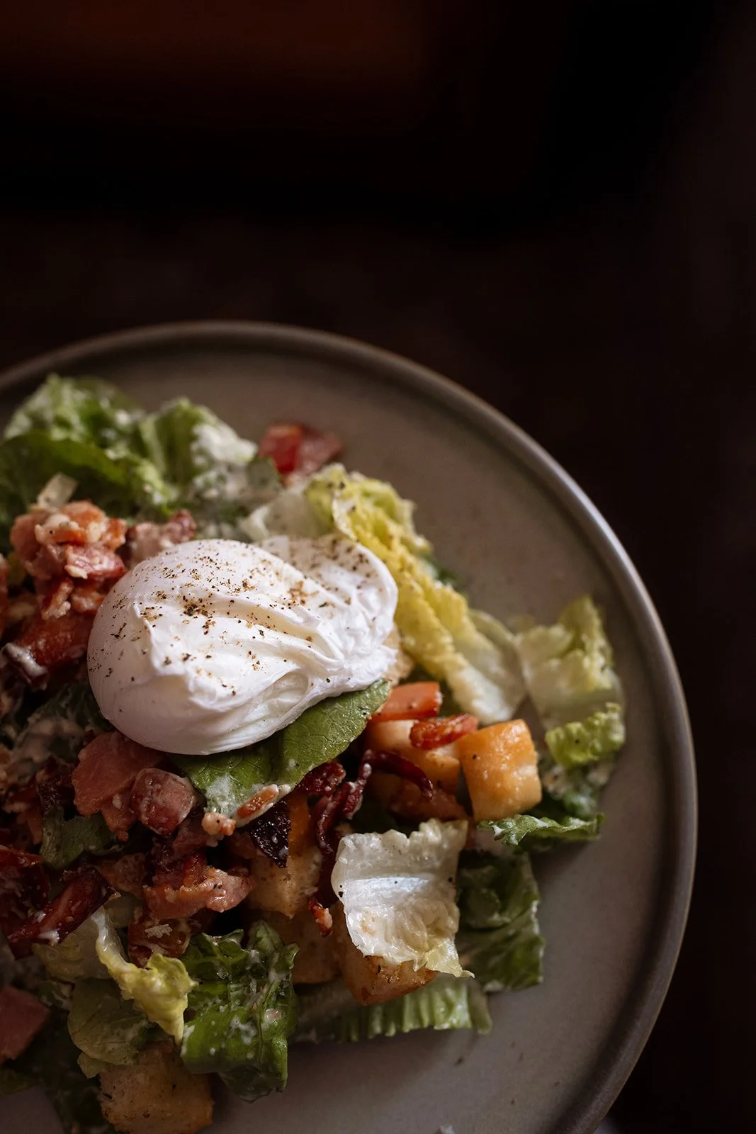 Close-up of a salad with bacon bits, croutons, lettuce, cherry tomatoes, a poached egg on top, and a light dressing in a gray bowl.