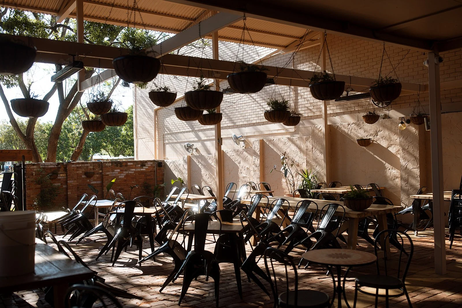 Empty outdoor patio with small tables and chairs, hanging pots with plants, and sunlight casting shadows on the brick floor.