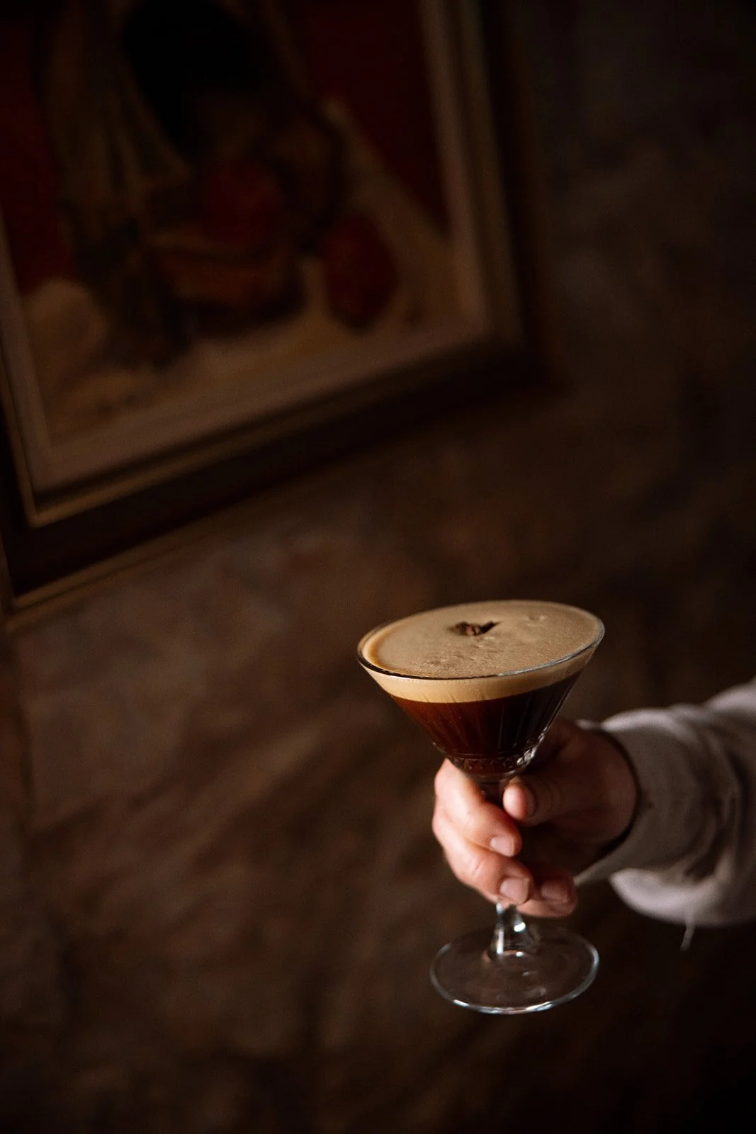 A person's hand holding a cocktail glass filled with a dark brown drink topped with foam, in front of a dark background with a framed picture.