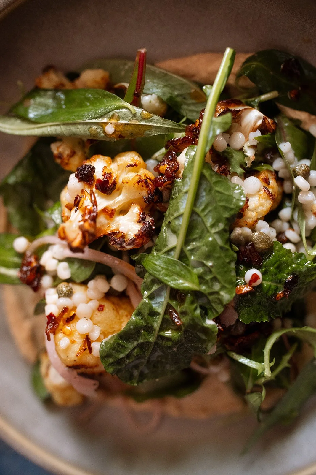 Close-up of a salad with grilled cauliflower, fresh greens, and small white crunchy grains, topped with sliced almonds.