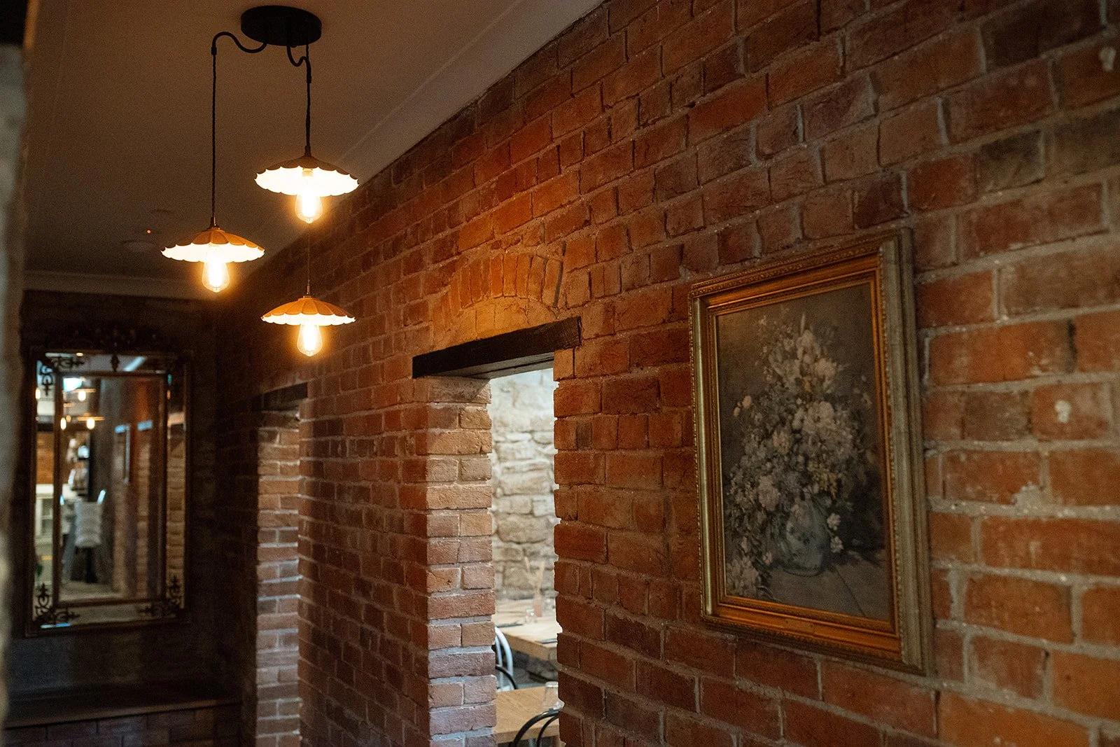 Interior view of a cozy room with red brick walls, hanging pendant lights, a framed painting of a flower bouquet, and a mirror reflecting part of the room.
