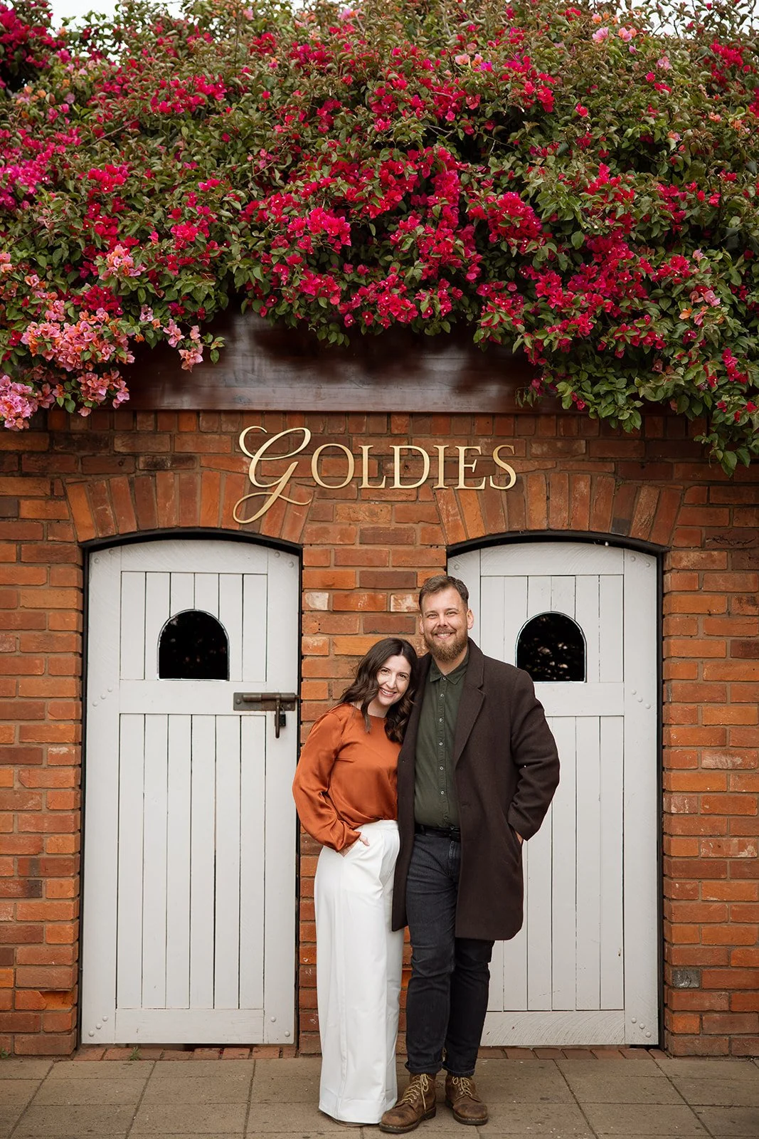 A smiling couple stands in front of a brick wall with white wooden doors labeled 'Goldies', surrounded by pink flowers.