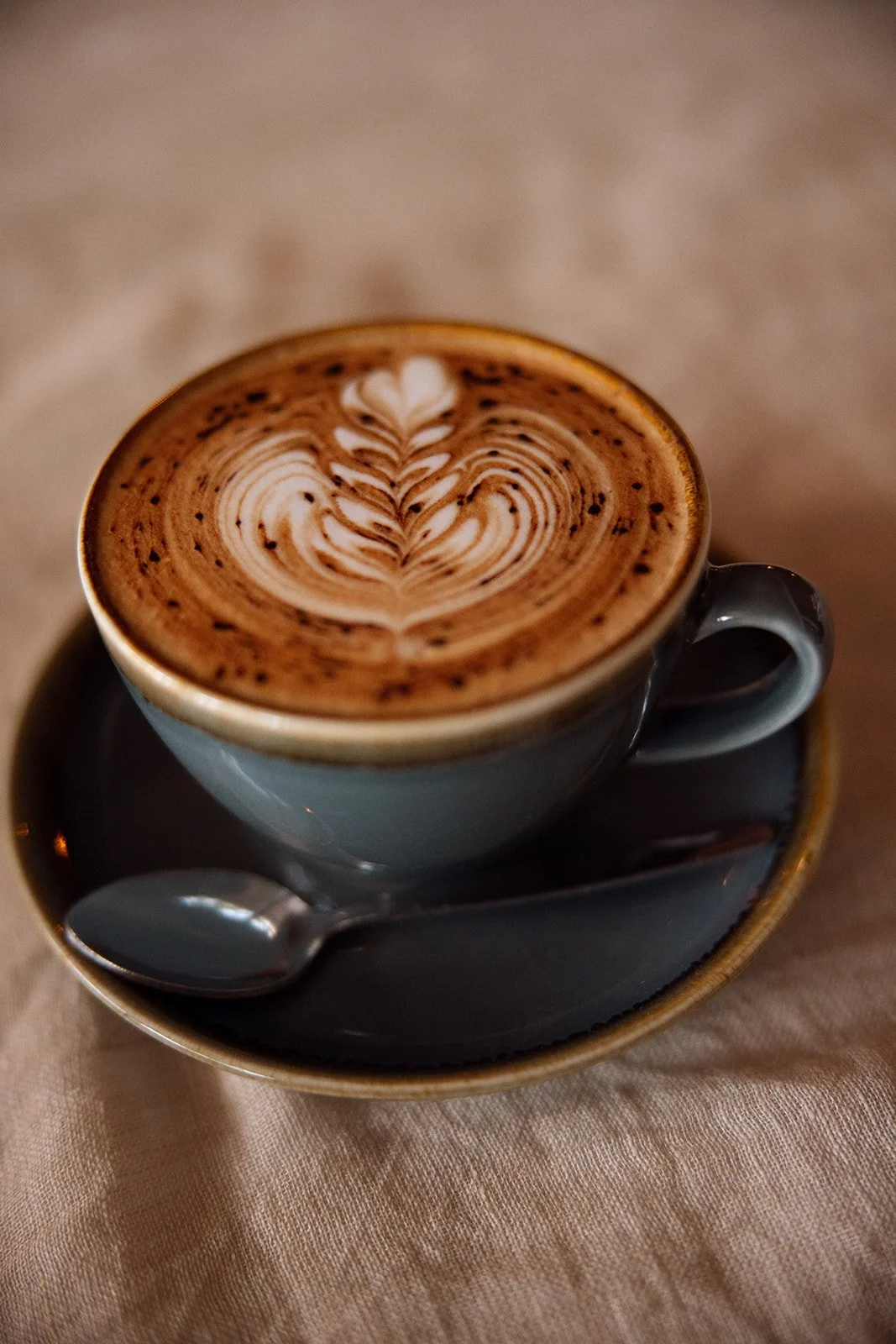 A cup of coffee with latte art on top, served on a saucer with a small spoon, placed on a beige tablecloth.