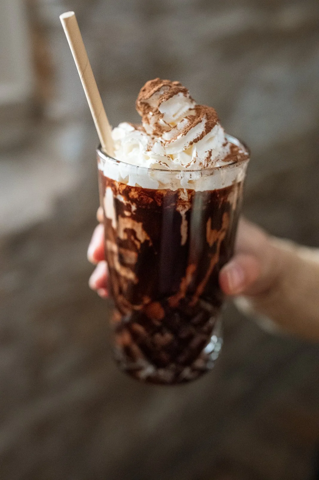 A hand holding a tall glass of chocolate milkshake topped with whipped cream and cocoa powder, with a drinking straw.