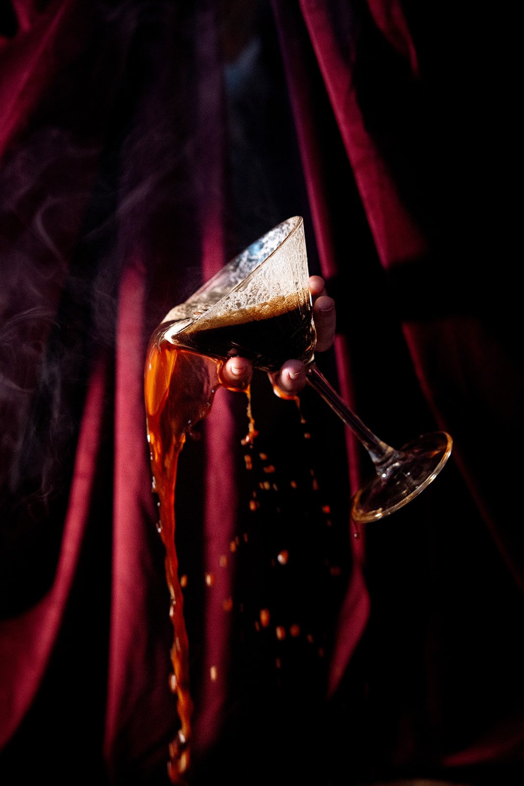 A person holding a tilted cocktail glass with a dark drink, with foam on top, against a dark background with red curtains.