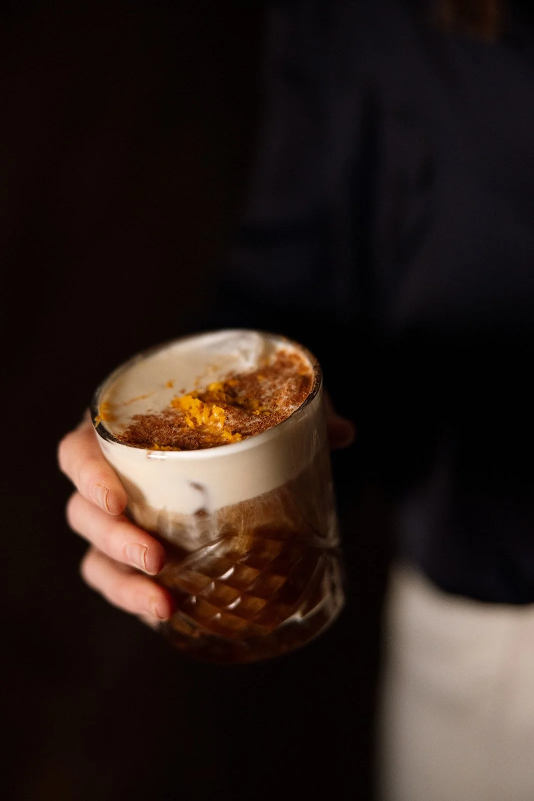 A person holding a glass of whipped coffee with a layer of cream and sprinkled cinnamon or cocoa powder on top.