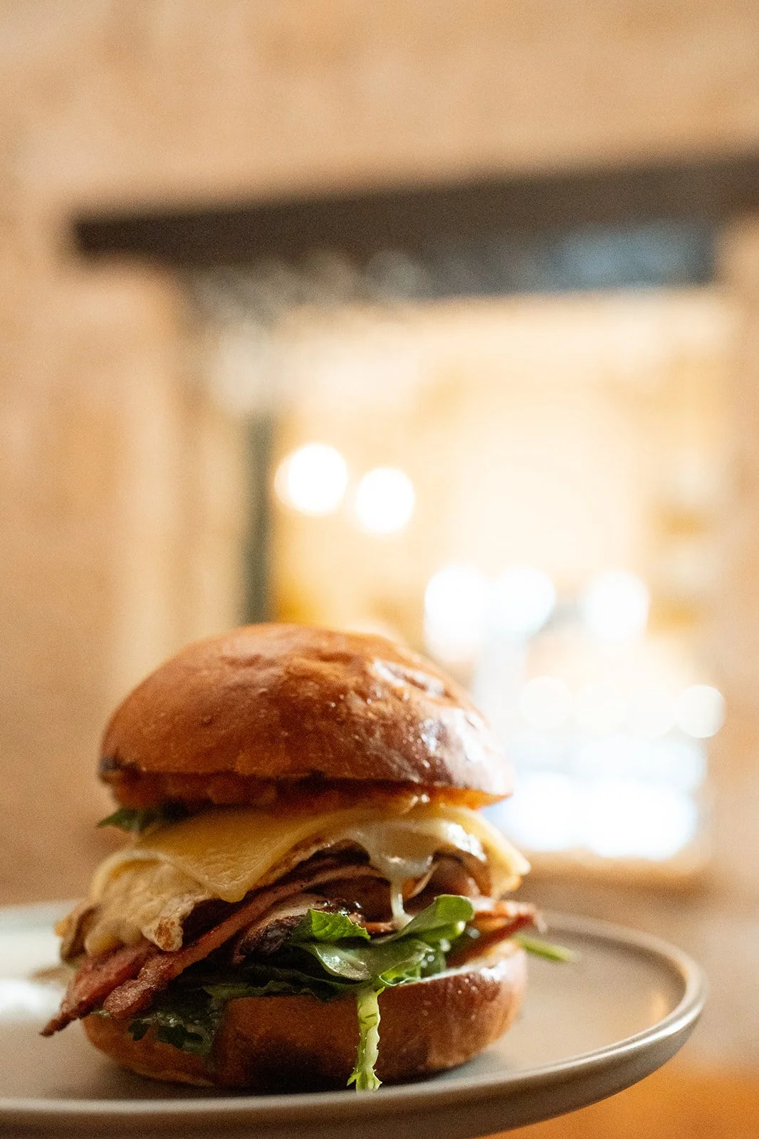 Close-up of a gourmet burger on a white plate, with lettuce, cheese, bacon, and a toasted bun, blurred warm indoor background.