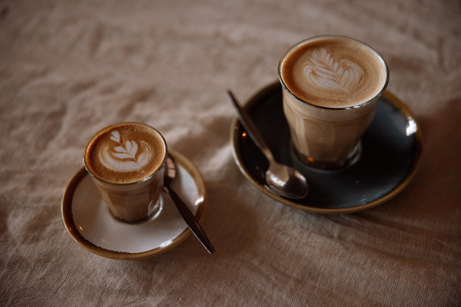 Two glasses of latte with latte art on top, placed on small cups with spoons on a wooden table.