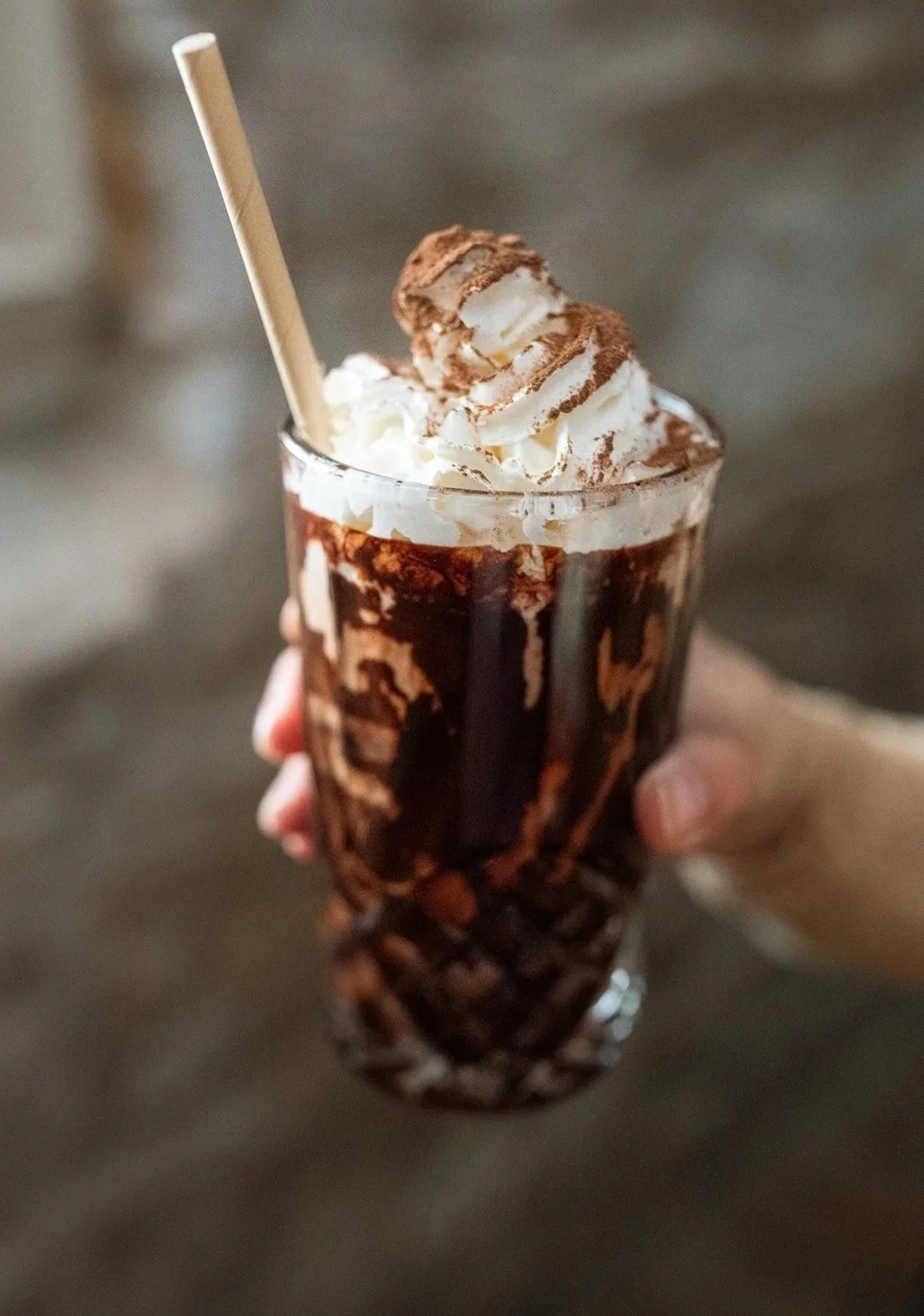 A hand holding a tall glass of iced coffee topped with whipped cream and dusted with cocoa powder, with a striped straw.