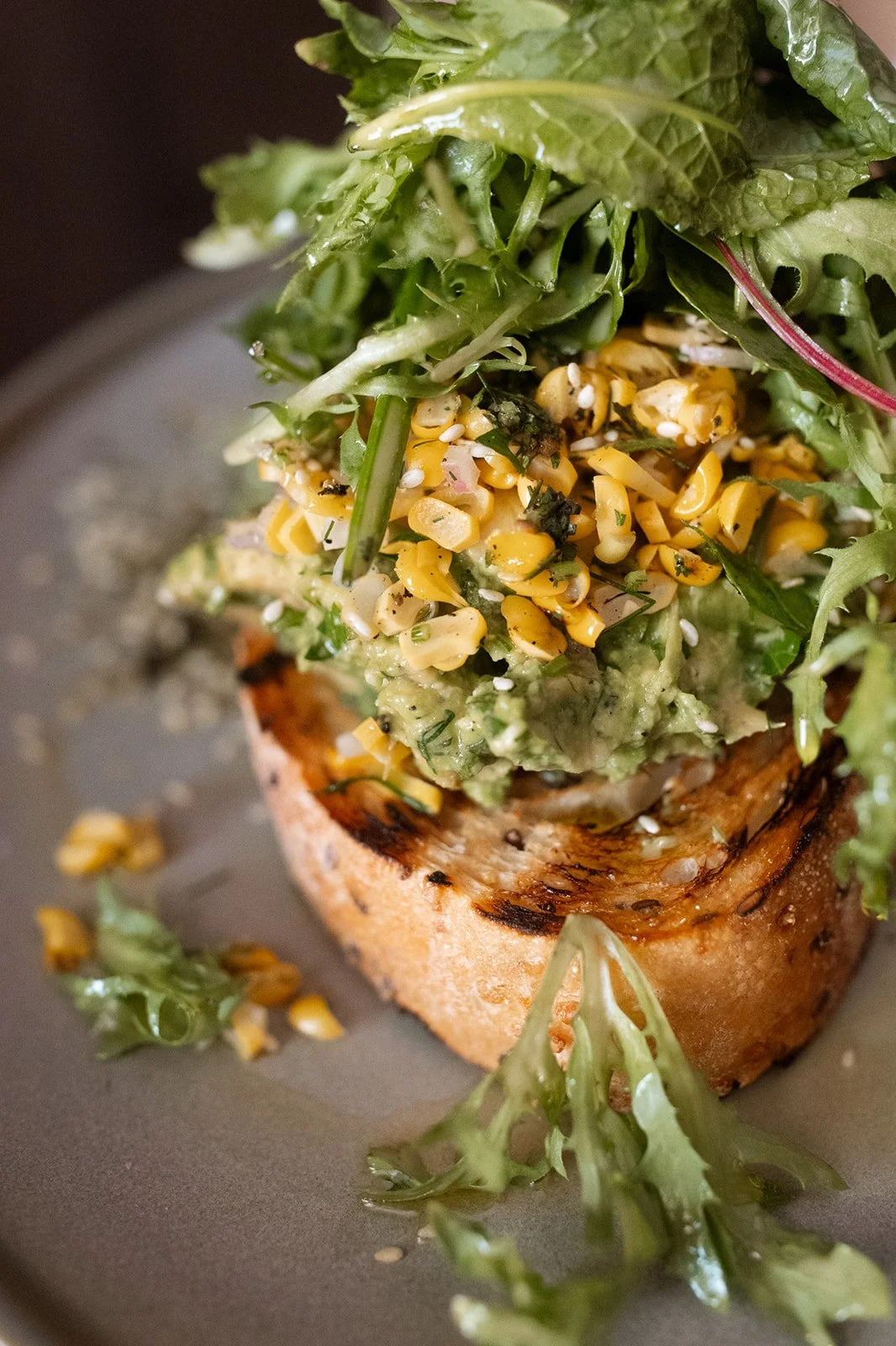 Close-up of a piece of toasted bread topped with guacamole, corn, and mixed greens, on a gray plate.