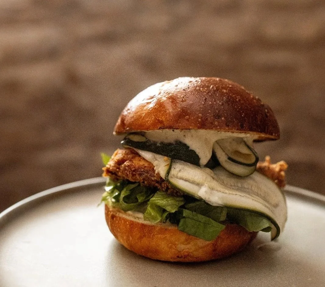 A close-up of a gourmet burger with a toasted bun, lettuce, fried chicken patty, a slice of fresh cucumber, a slice of pepper, and a creamy sauce, served on a white plate.